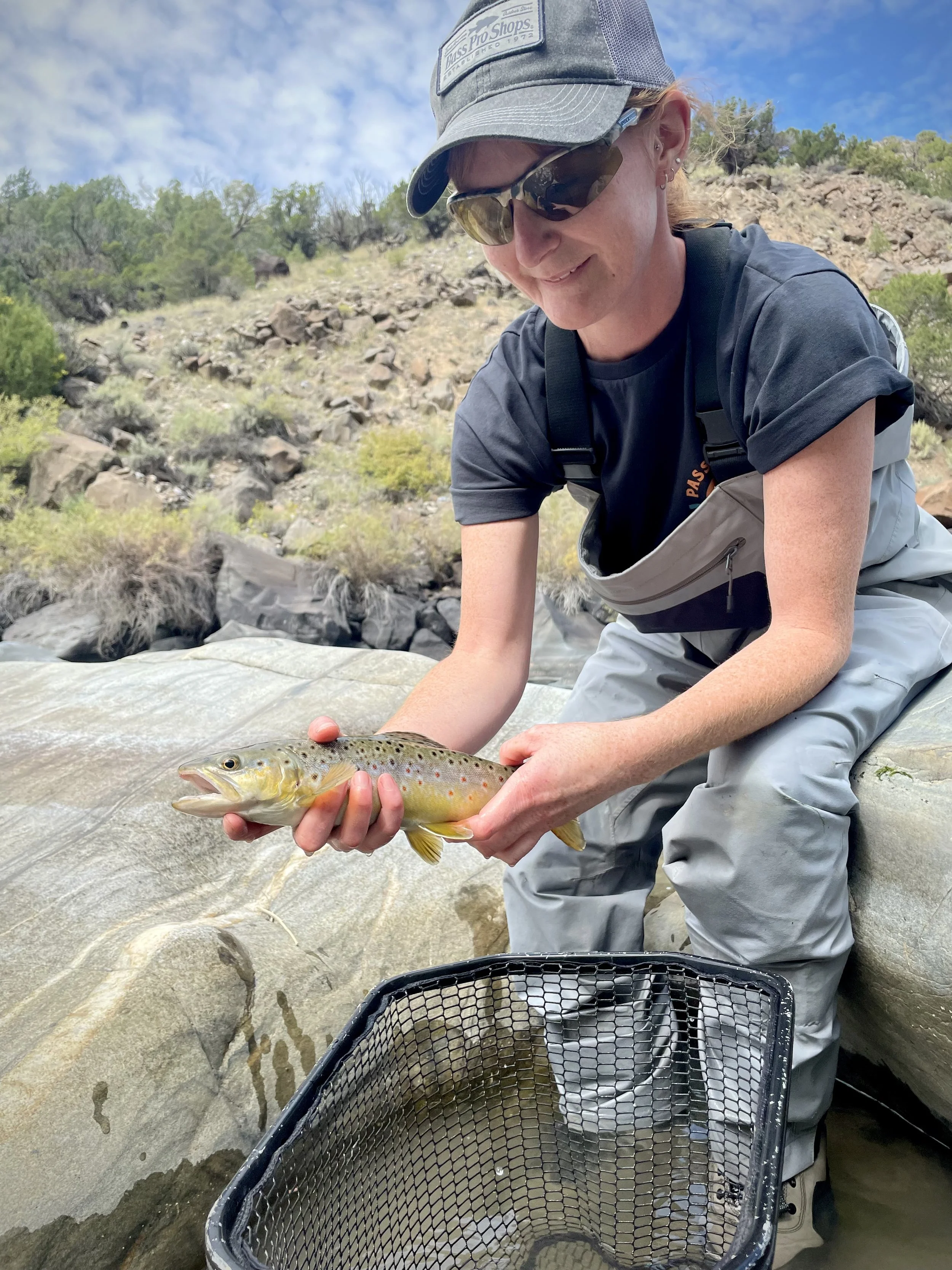 A woman holding a small rainbow trout fish outdoors near a river, with a fishing net below, rocky terrain, and green trees in the background.