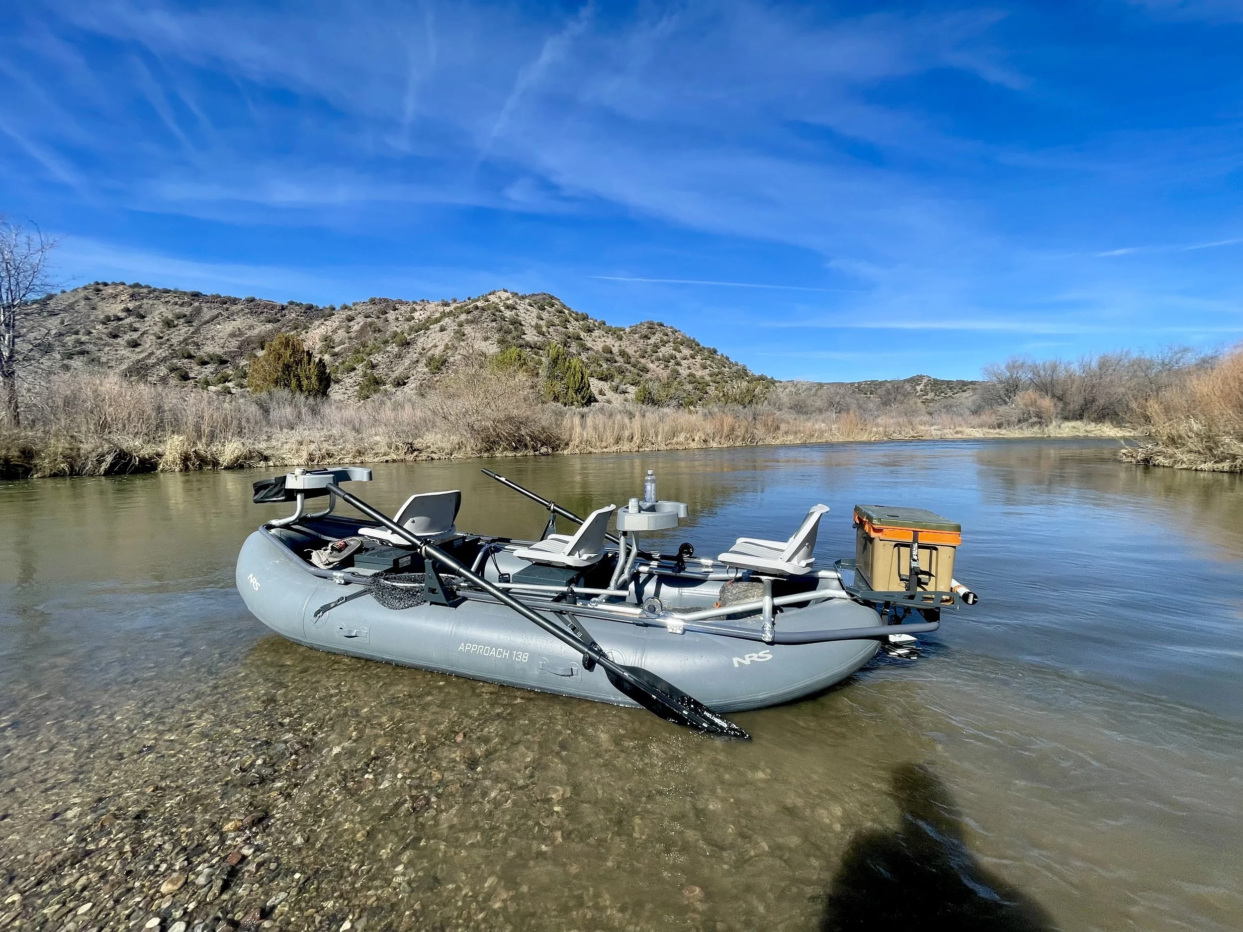 A gray inflatable boat with seats, paddles, and a cooler on a calm river with hills and blue sky in the background.