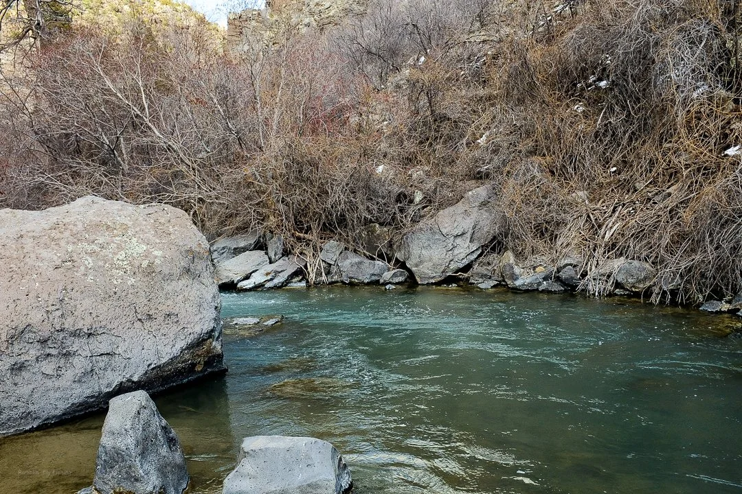 A peaceful river flowing through a rocky and wooded landscape with leafless bushes and trees.