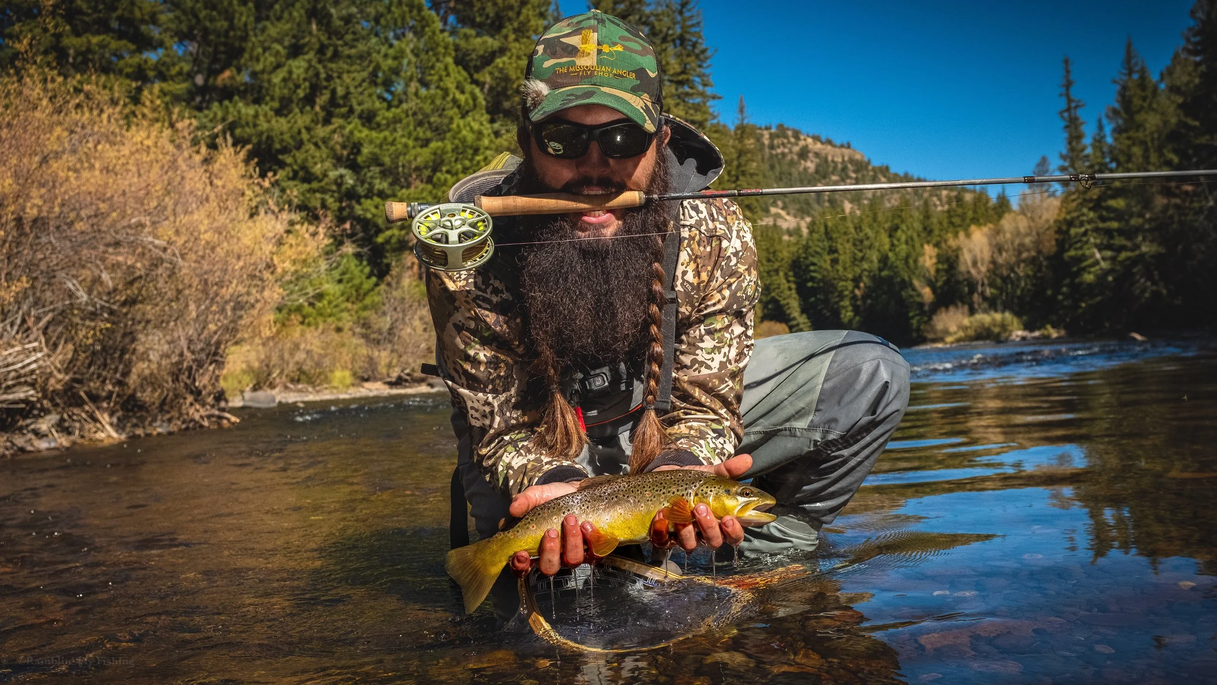 A man with sunglasses and a patterned camouflage jacket kneels in a river, holding a freshly caught brown trout with both hands, with a fishing rod resting on his shoulder, surrounded by forested landscape under a clear blue sky.