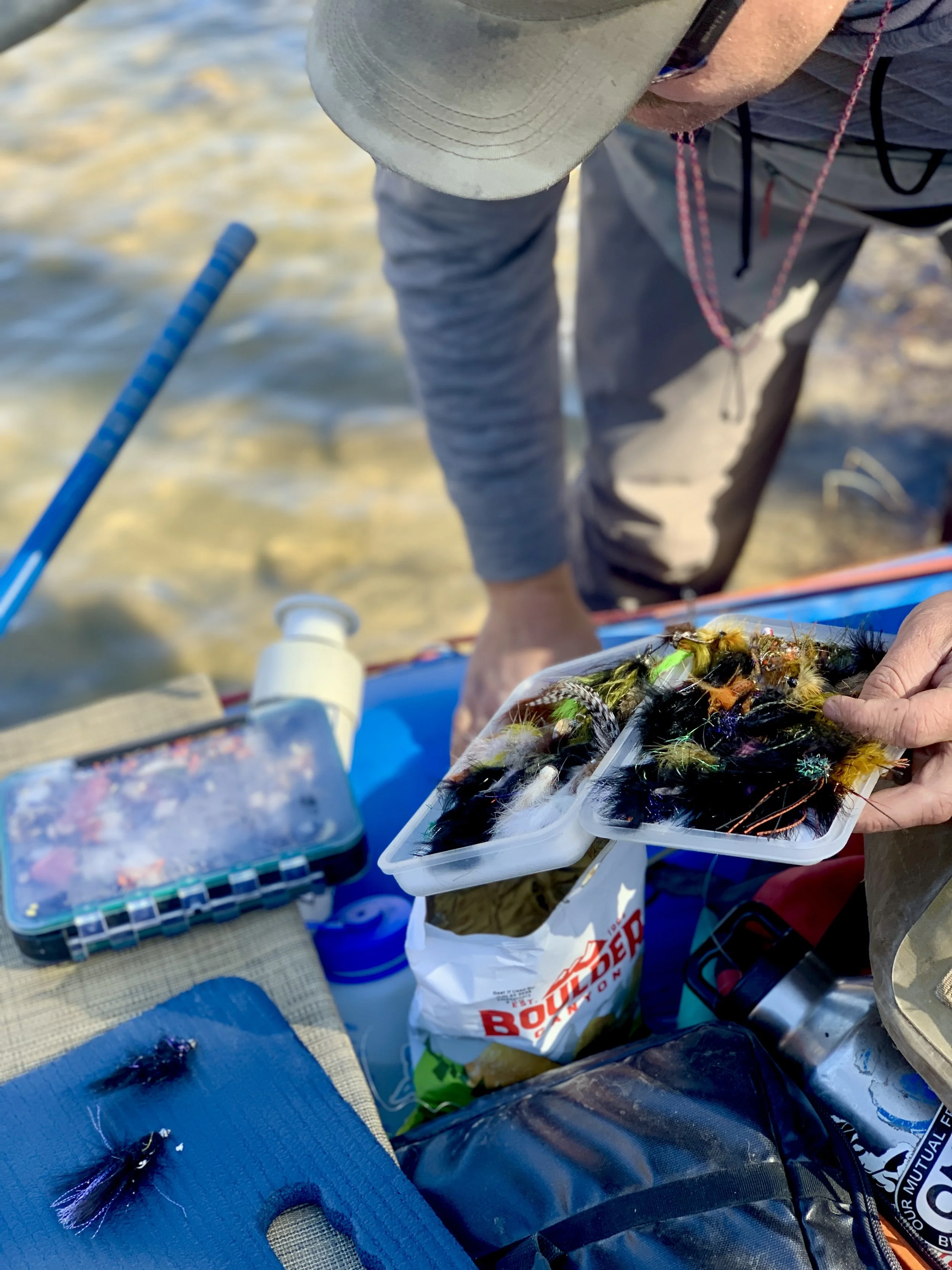 Person holding containers of colorful fishing flies on a boat near water, with fishing gear and supplies on the surface.