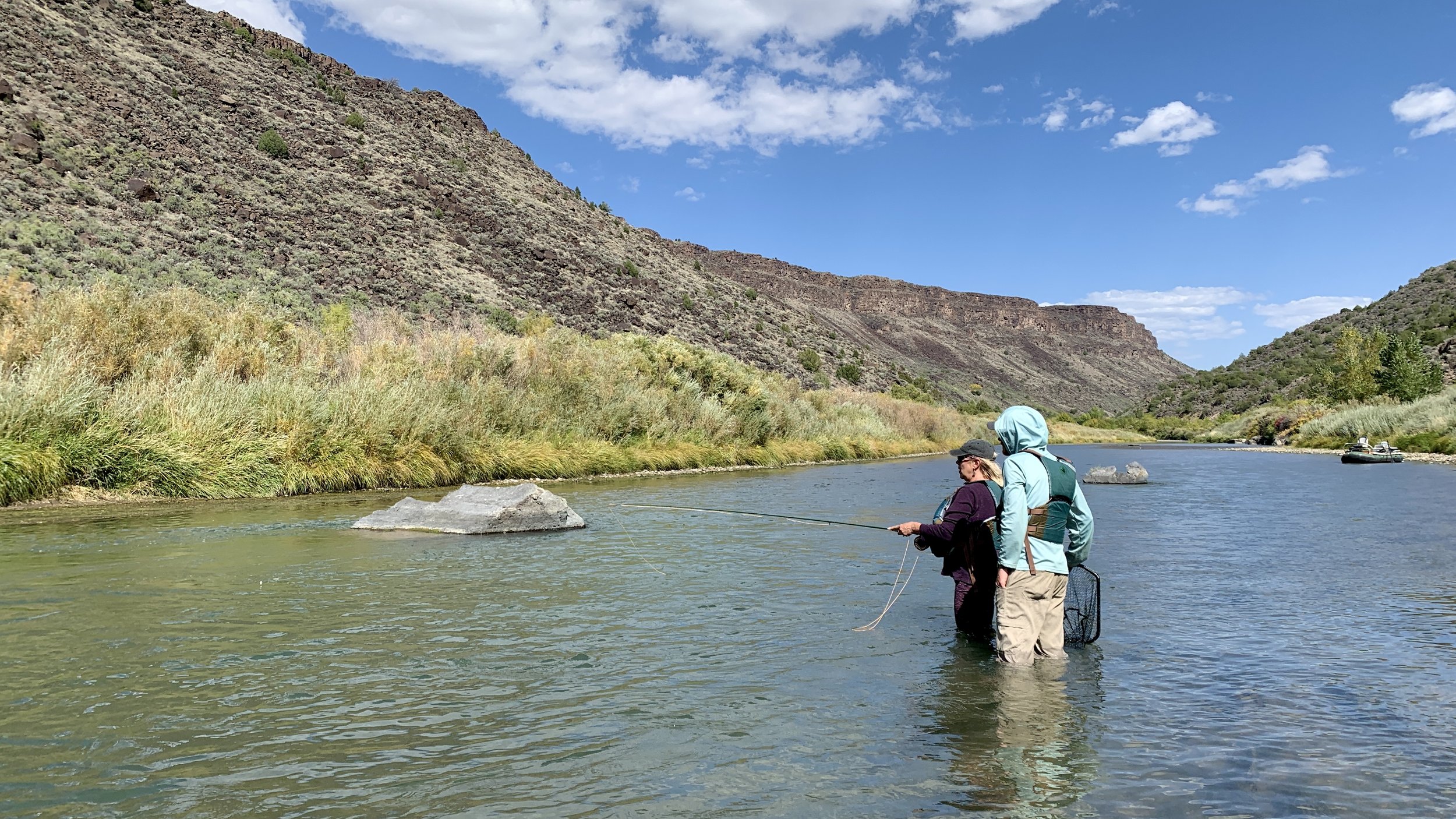 Two people fishing in a shallow river surrounded by mountains and green vegetation under a partly cloudy sky.