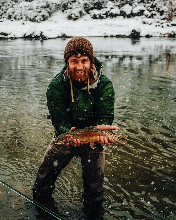 A man with a beard, wearing a brown knit hat and a green waterproof jacket, is standing in a river holding a fish he caught. Snow is falling and snow covers the ground and the riverbank in the background.