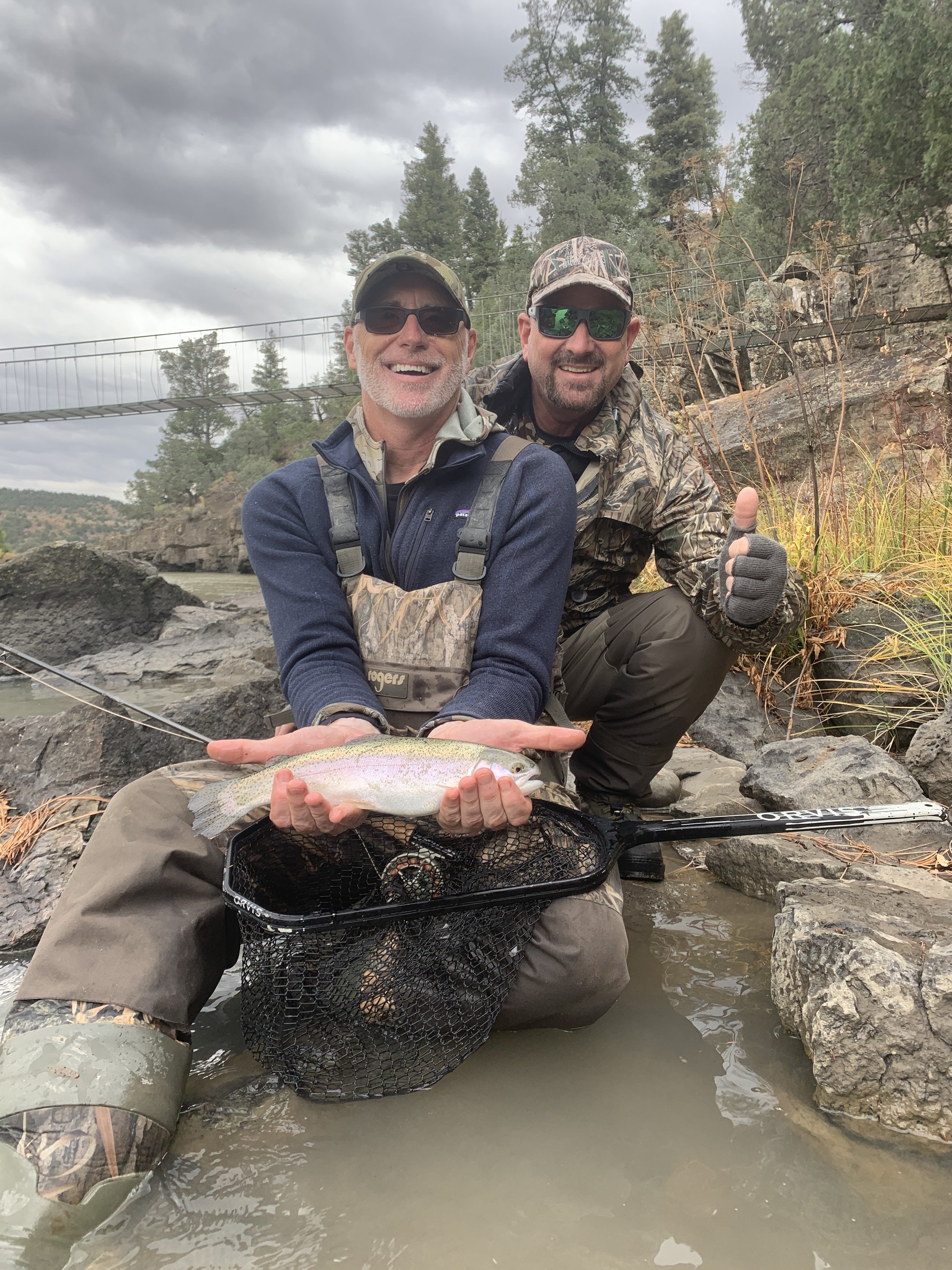 Two men in outdoor gear kneeling in a river, holding a fish with one man holding a fishing net. They are smiling, and one is giving a thumbs-up. The background shows trees, rocks, and a suspension bridge with cloudy skies overhead.