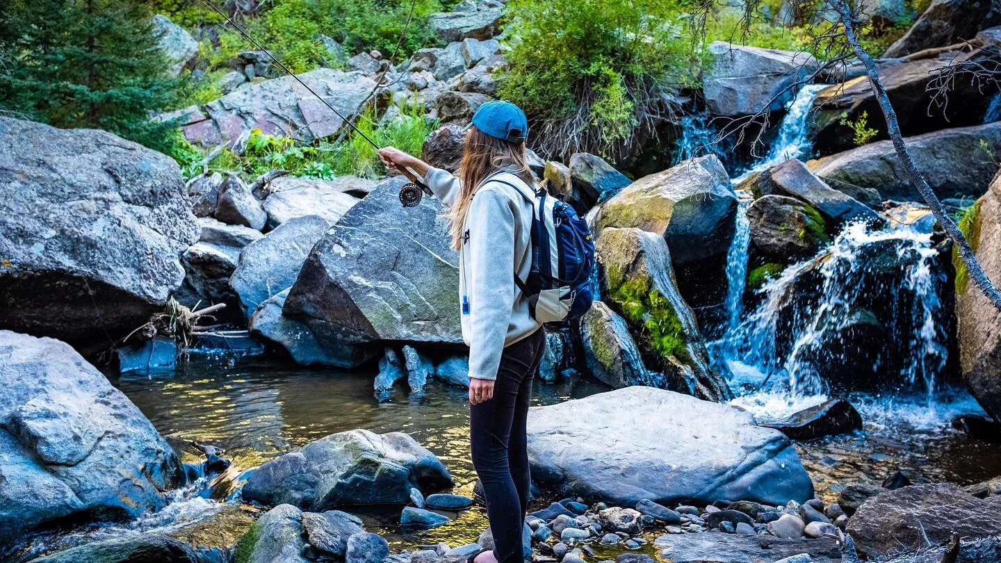 A woman with a backpack fishing in a rocky creek surrounded by trees with a small waterfall in the background.