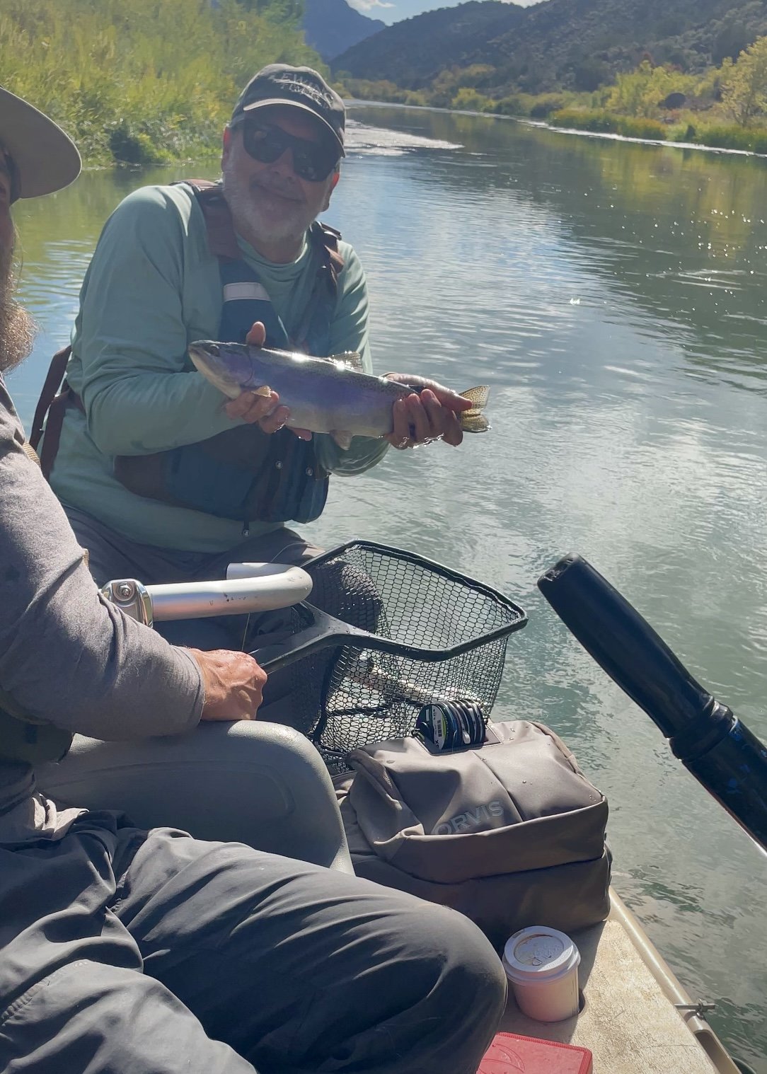A man in sunglasses and a cap holding a fish on a boat by a river with mountains in the background.