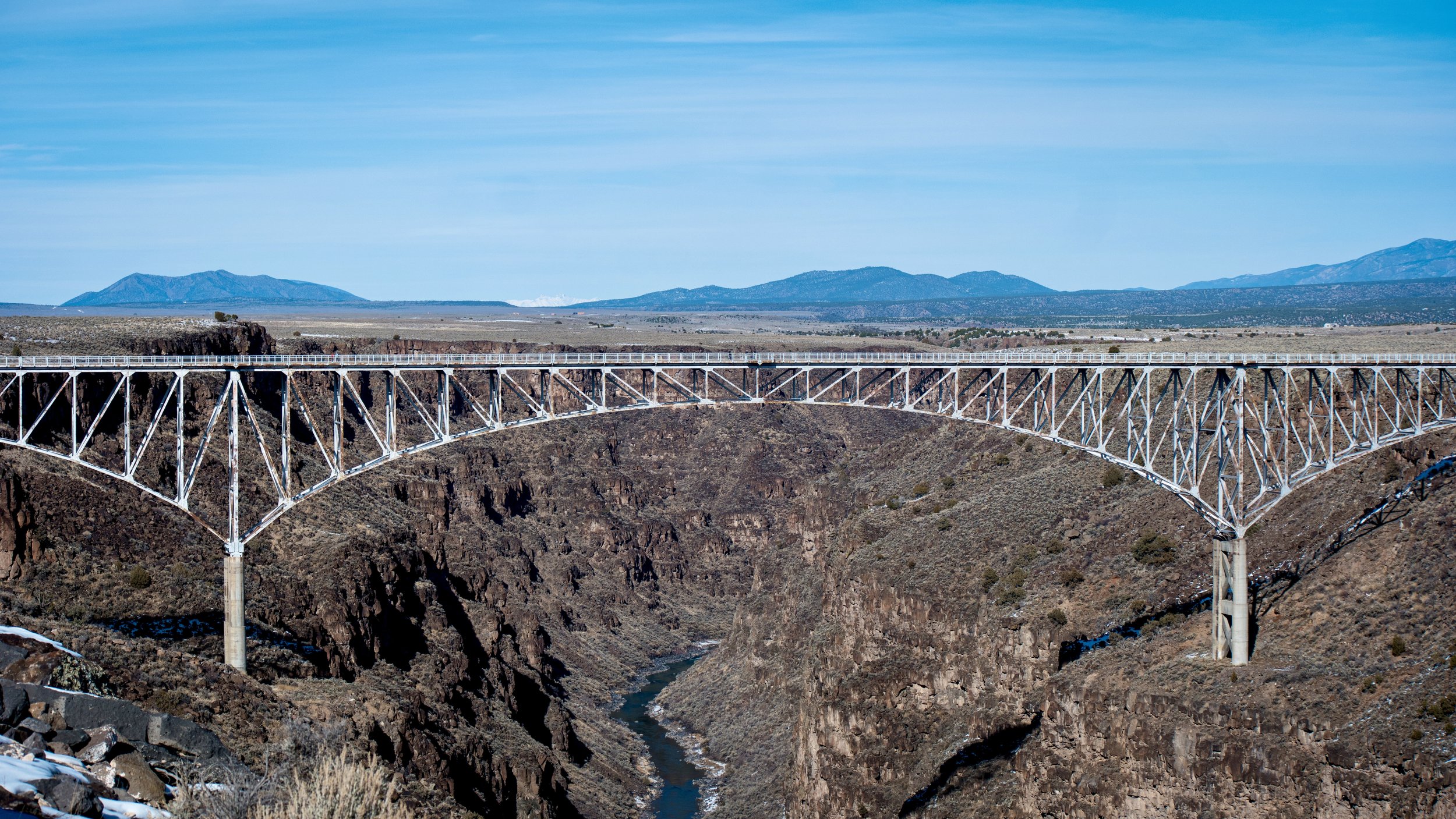 Steel arch bridge spanning a rocky canyon with a river at the bottom, under a blue sky with distant mountains.