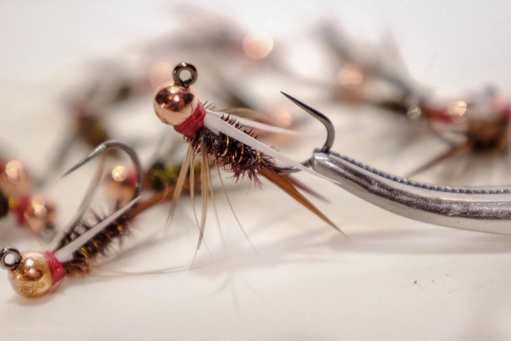 Close-up of a fishing fly connected to a hook, with blurred fishing lures in the background.