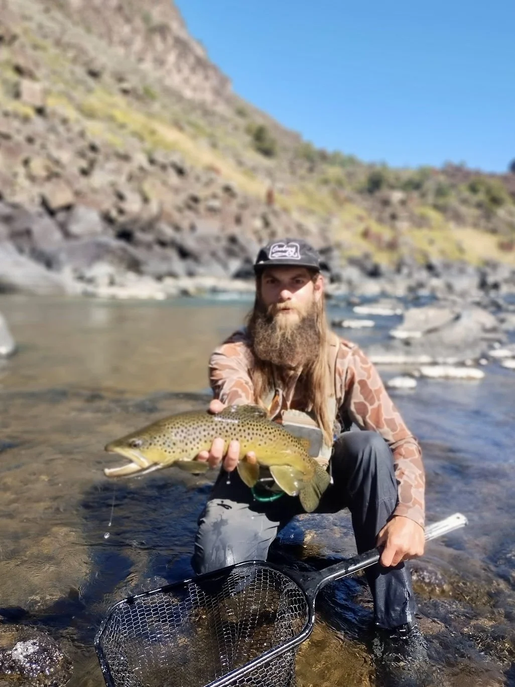 Man in camouflage jacket and black cap kneeling in river, holding a large trout with a fishing net nearby, rocky terrain with slope and blue sky in the background.