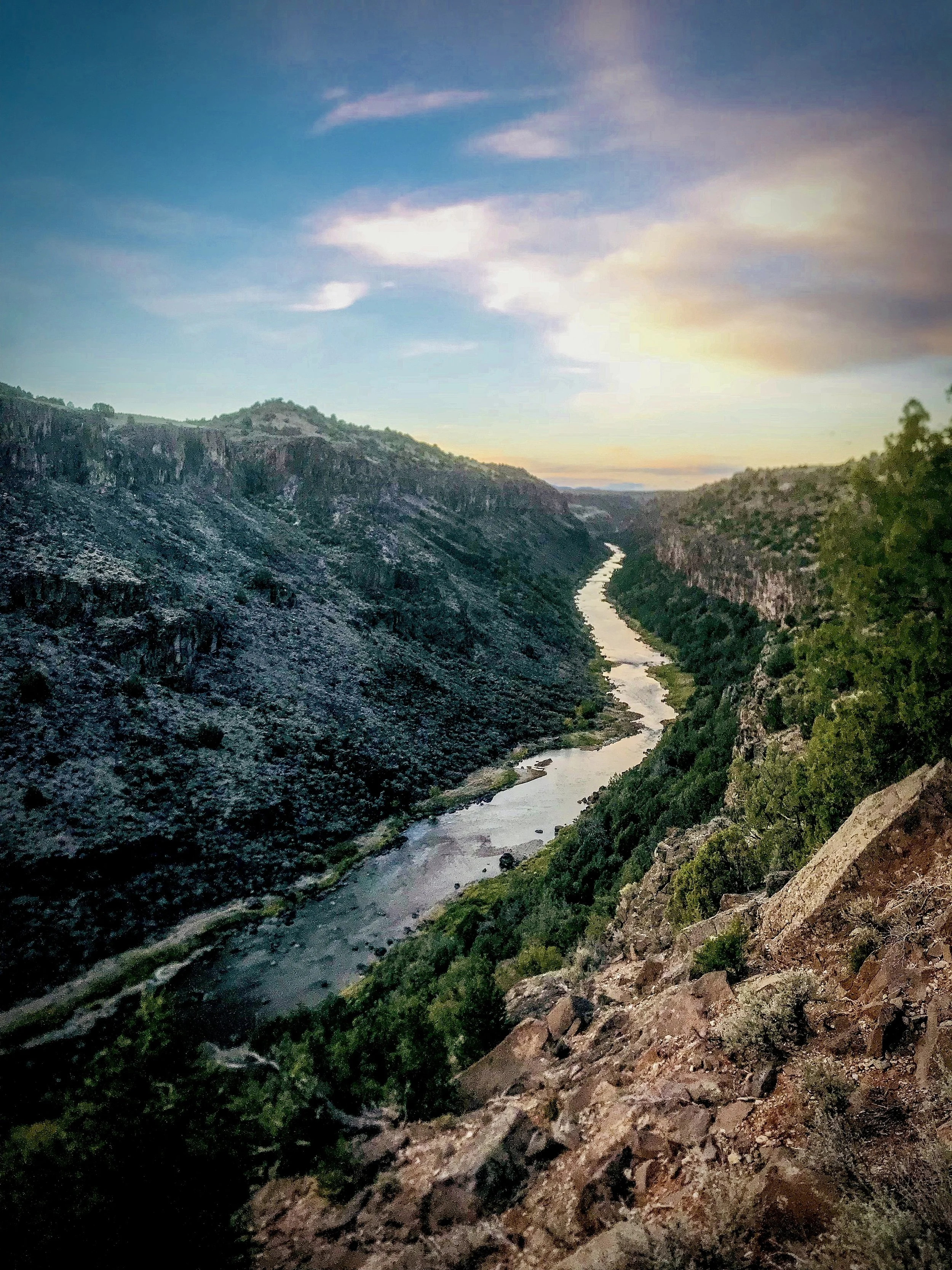 A river runs through a deep canyon with rugged walls, green trees on the sides, and a partly cloudy sky above during sunset.