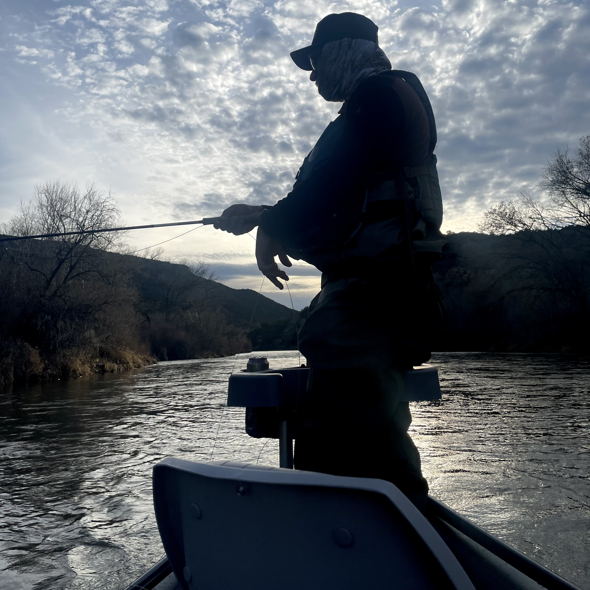 Silhouette of a person fishing on a boat on a river, with mountains and trees in the background under a cloudy sky.