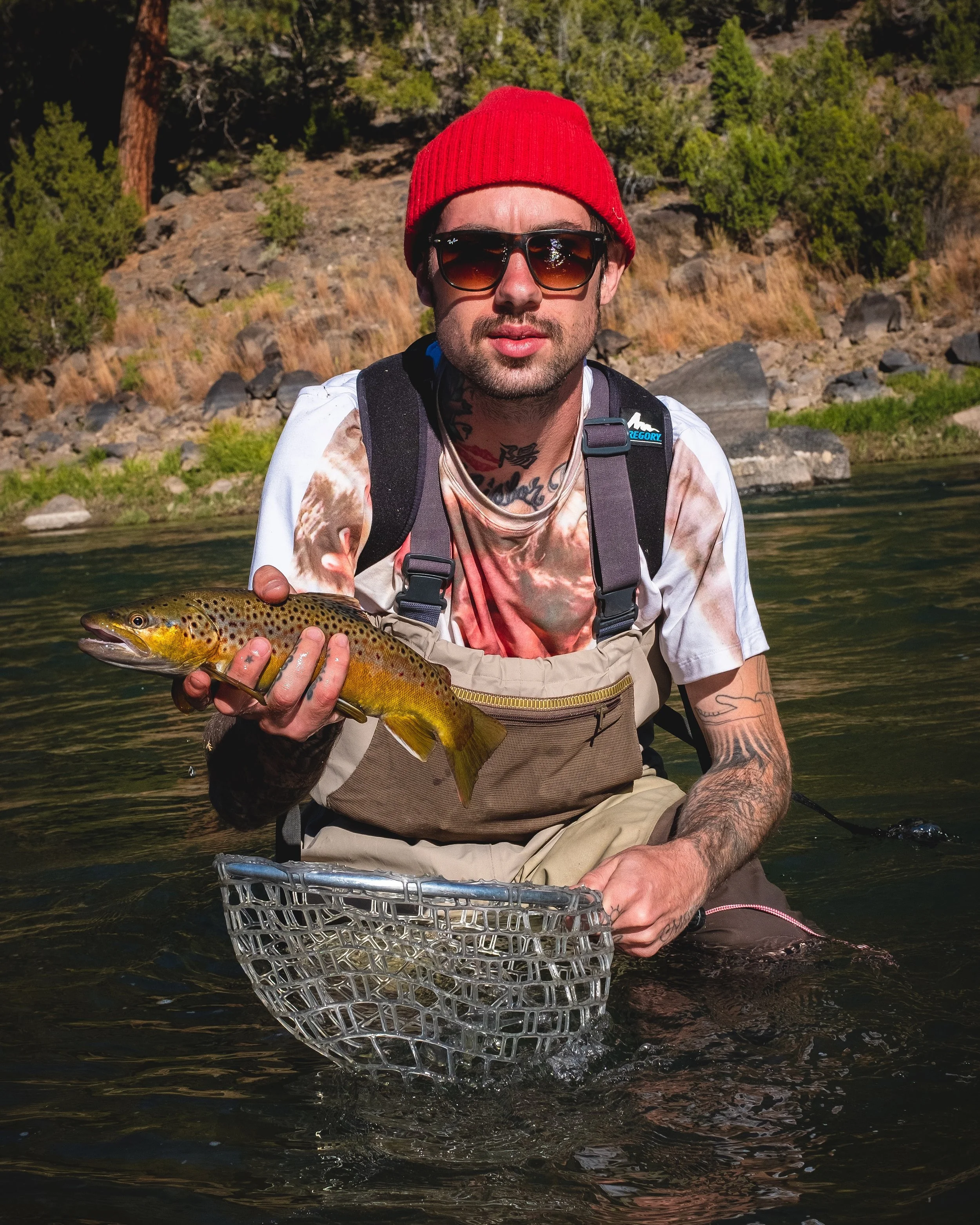 Man wearing a red beanie and sunglasses holding a freshly caught brown trout in a river, with a forested hillside in the background.