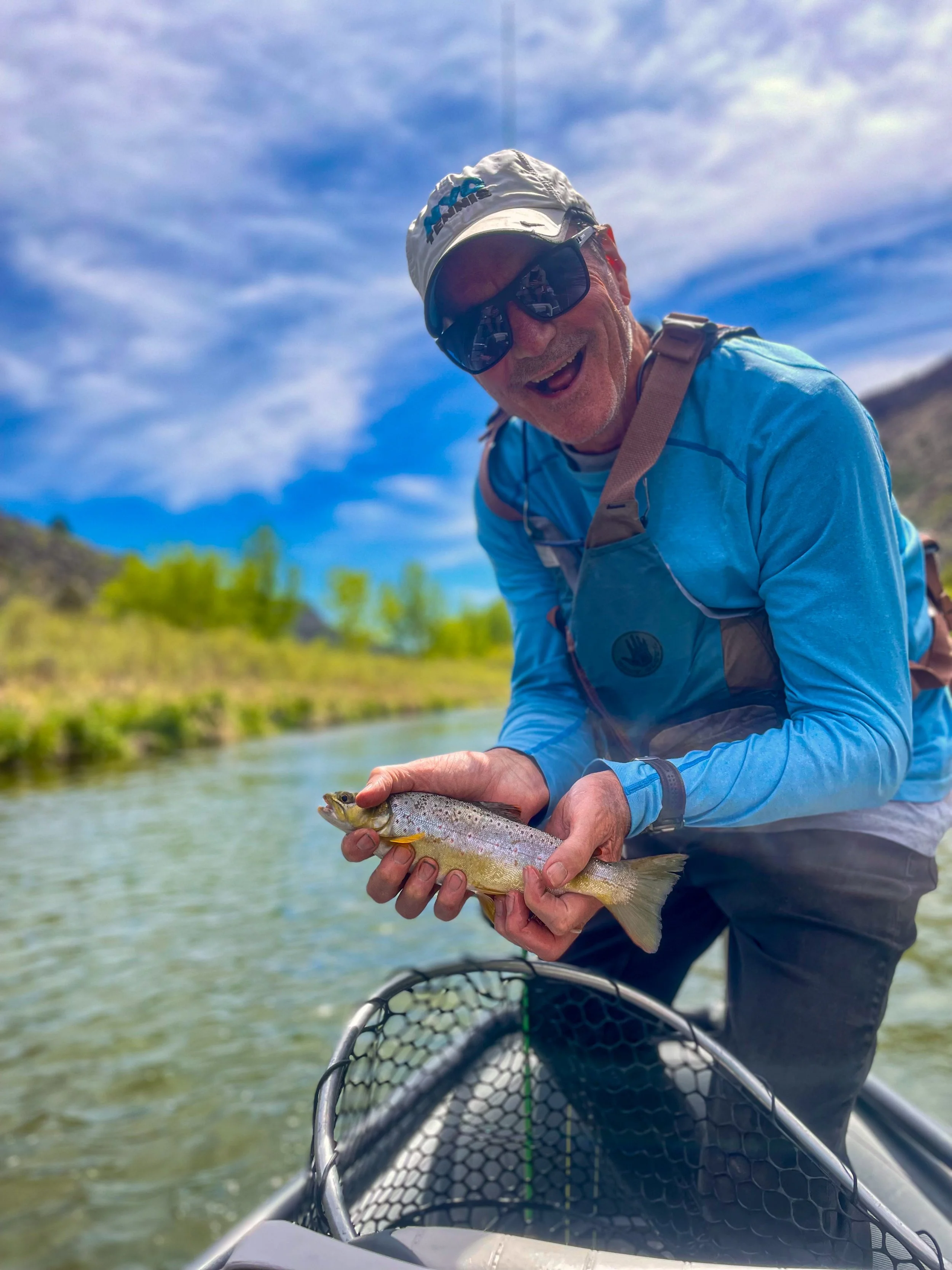A man wearing sunglasses, a white hat, and outdoor clothing is holding a fish in his hands near a river or lake. He appears to be smiling and enjoying a fishing trip with a fishing net in front of him. The background shows a blue sky with scattered clouds and green trees.