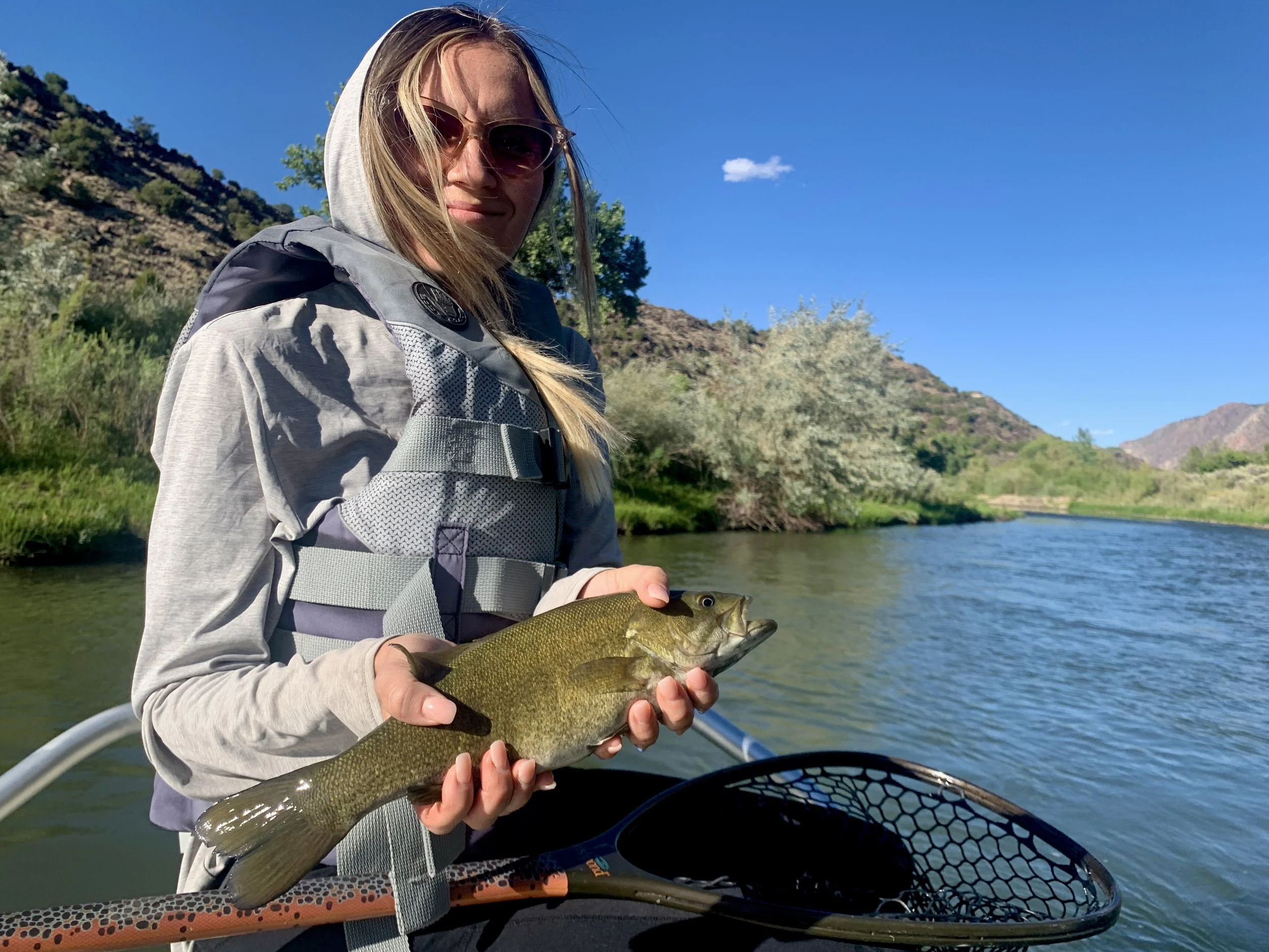 A woman wearing sunglasses, a gray hooded jacket, and a life vest holds a fish with the river and green hills in the background.