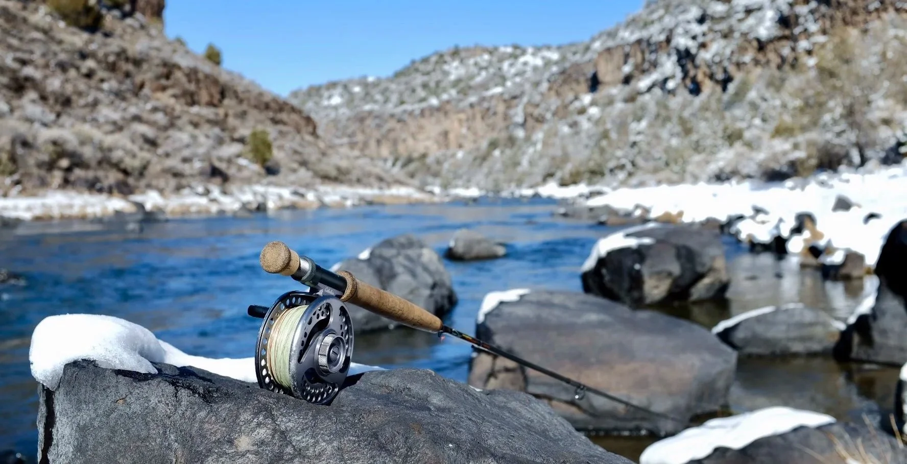 Fishing rod placed on a snow-covered rock near a river with rocks, mountains, and snow in the background.