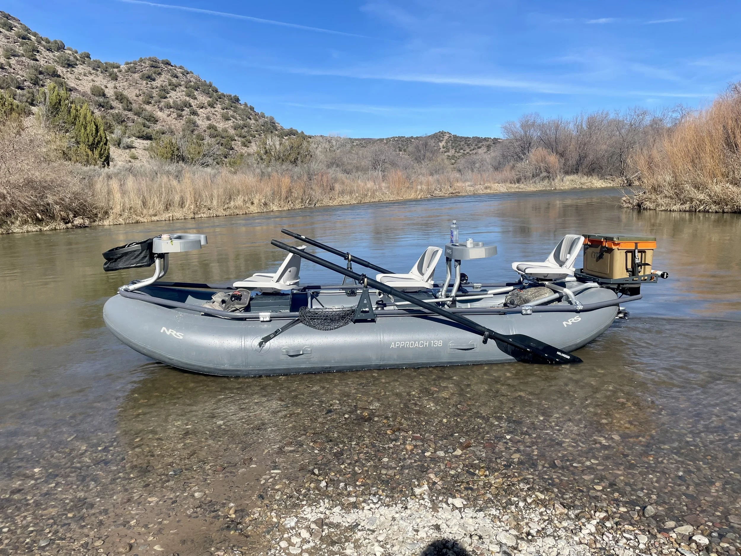 Inflatable fishing boat on a river with fishing rods, chairs, water bottles, and a storage box, set against a natural landscape with mountains, trees, and a blue sky.