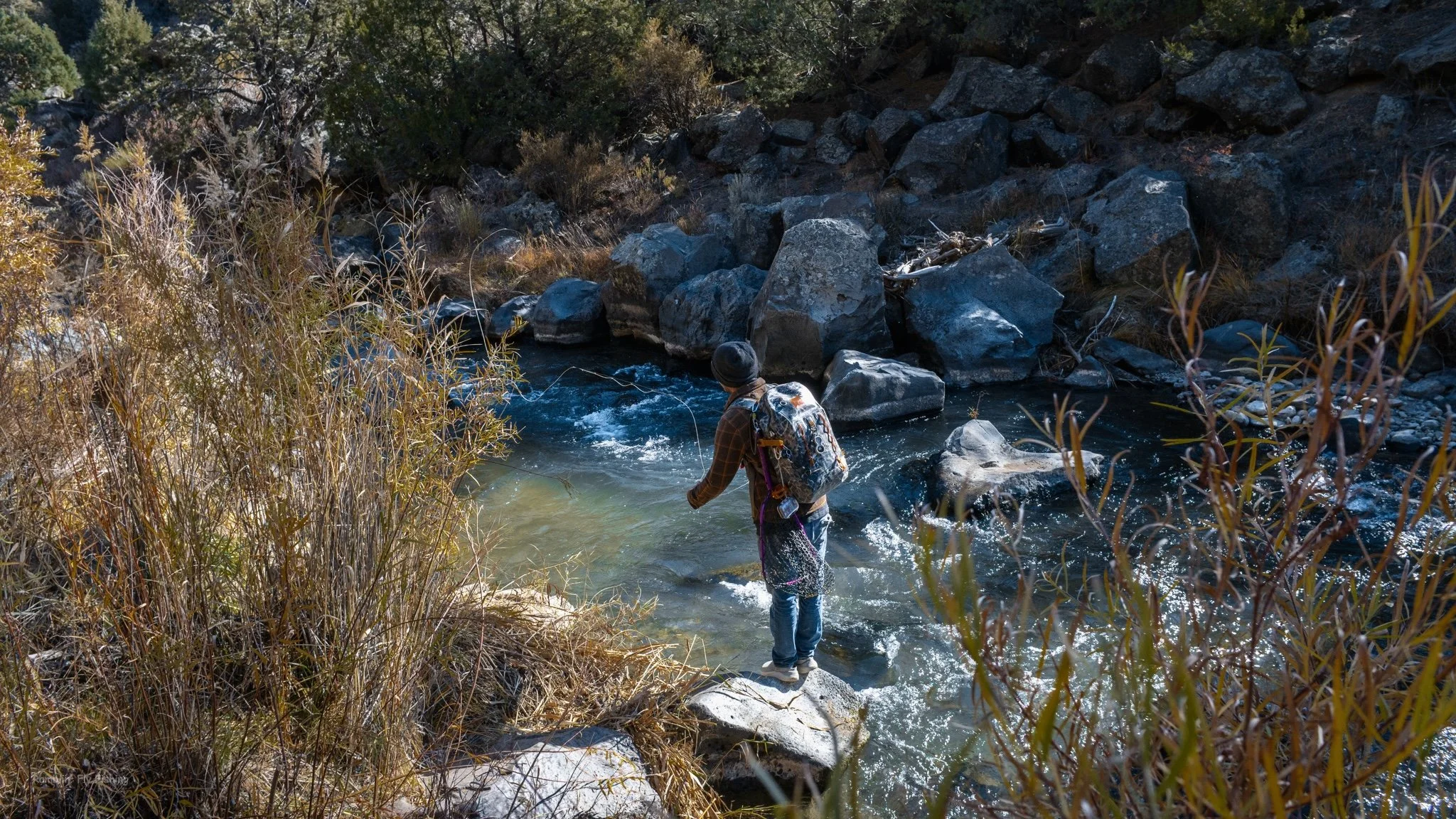 Person fishing in a rocky river surrounded by autumn foliage and trees.