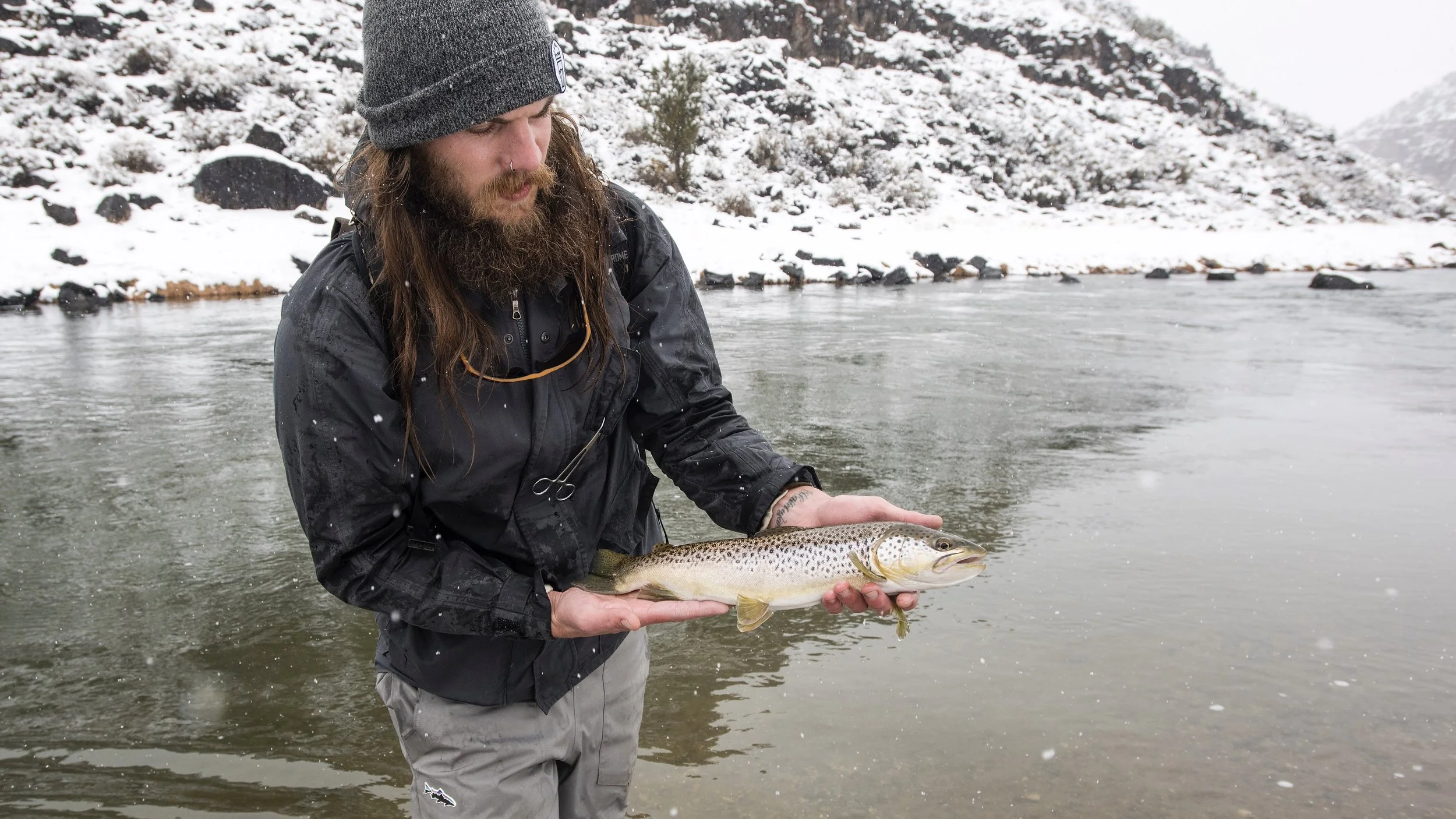 A man standing in a river holding a large fish with snow-covered rocks and trees in the background.