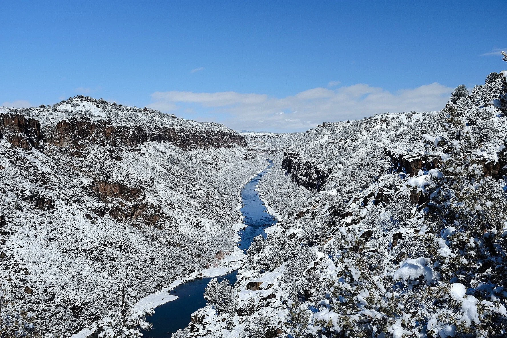 Snow-covered canyon with a river running through it and snow-laden trees against a blue sky.
