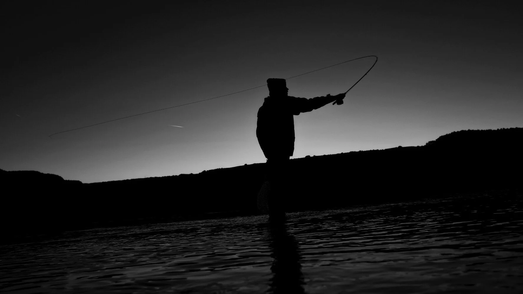 Silhouette of a person fishing in water at dusk or dawn, with a hill in the background.
