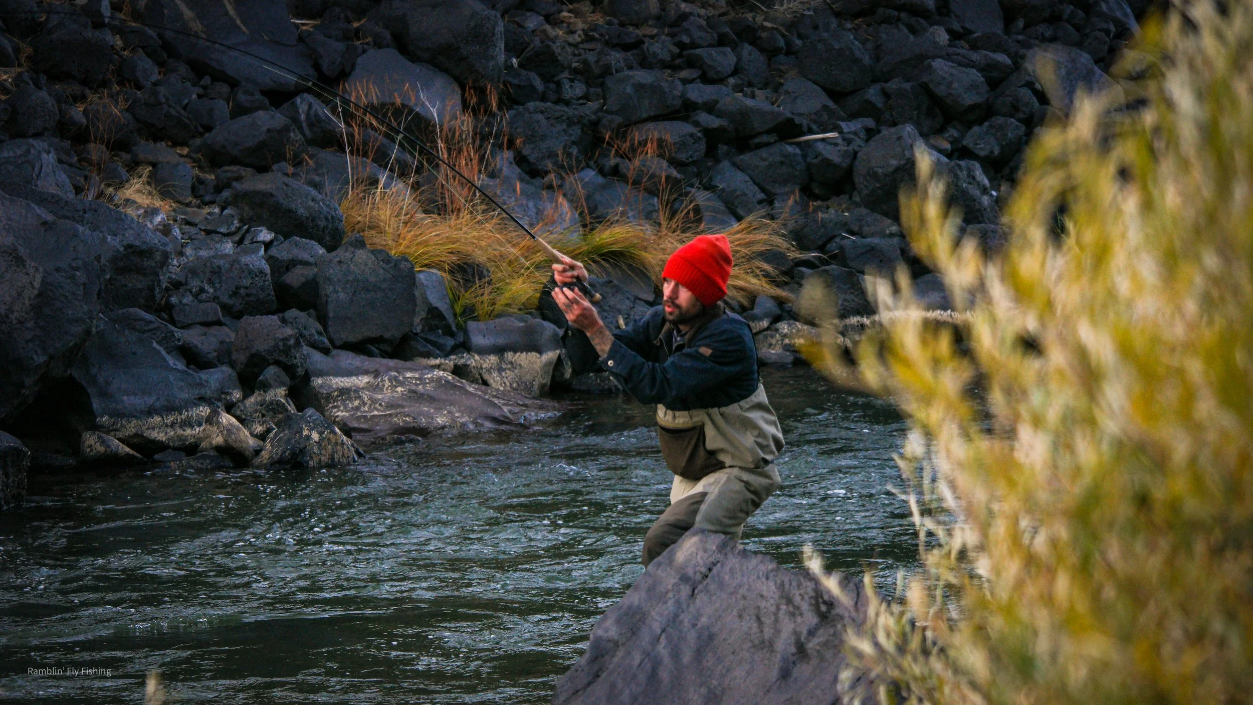 A man wearing a red beanie and waders fishing in a river with rocky banks and some yellowish-green vegetation.