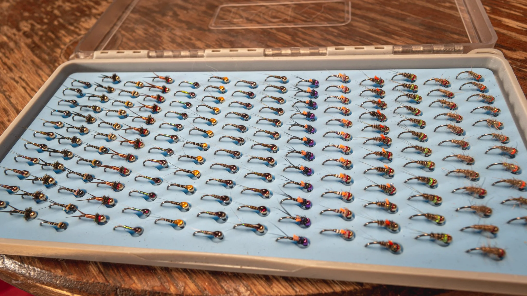 A plastic box filled with numerous small, colorful fishing flies arranged in rows on a blue foam insert.