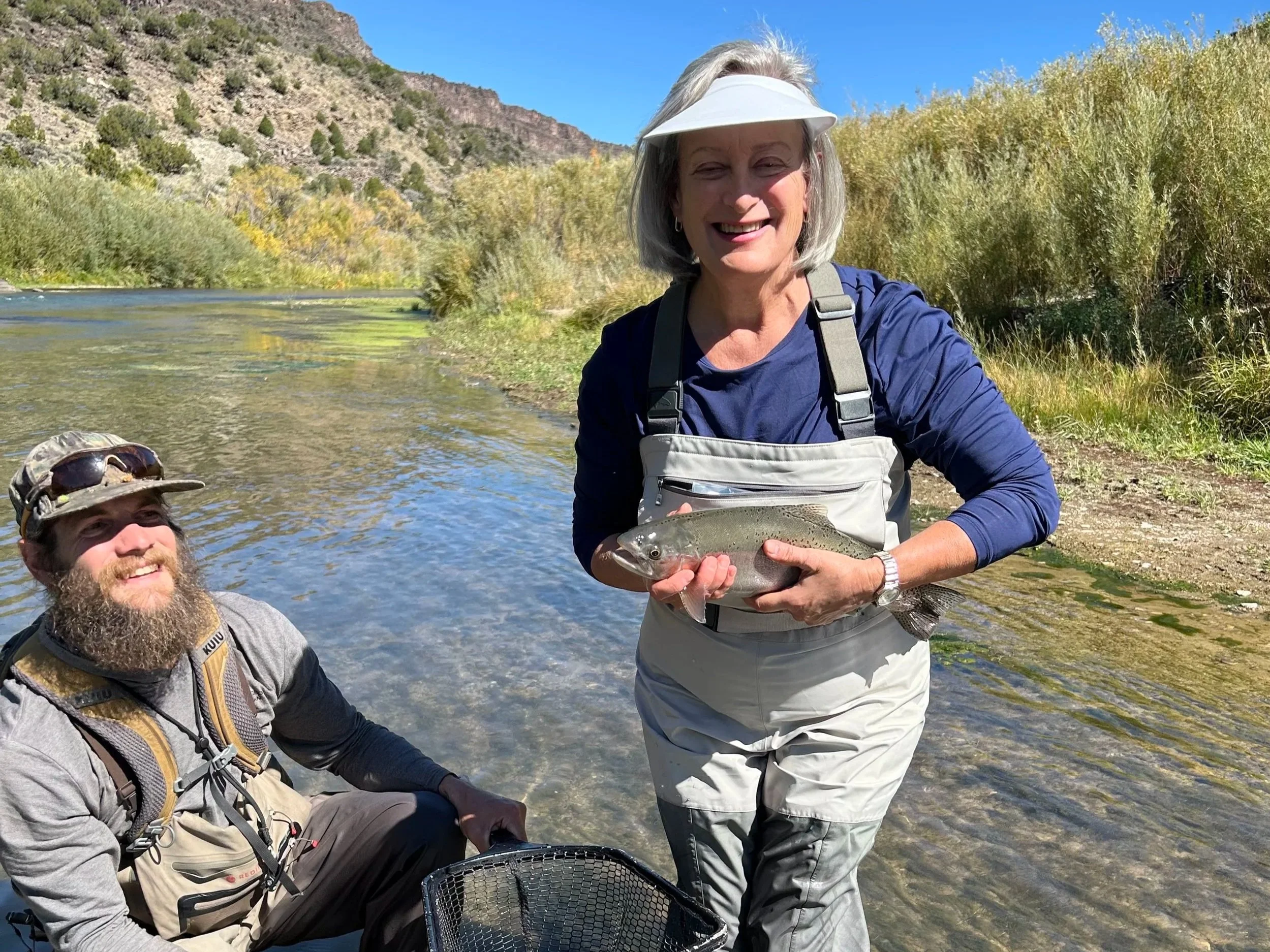 A smiling woman holding a fish she caught while fishing in a river, with a man sitting in the water nearby, both in outdoor fishing gear, surrounded by trees and mountains under a clear blue sky.