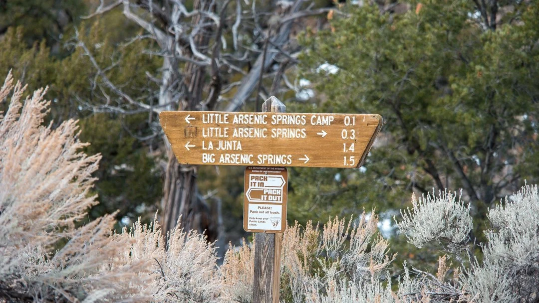 Wooden trail sign indicating directions and distances to Little Arsenic Springs Camp, Little Arsenic Springs, La Junta, and Big Arsenic Springs, with a smaller instruction sign below about trash disposal.