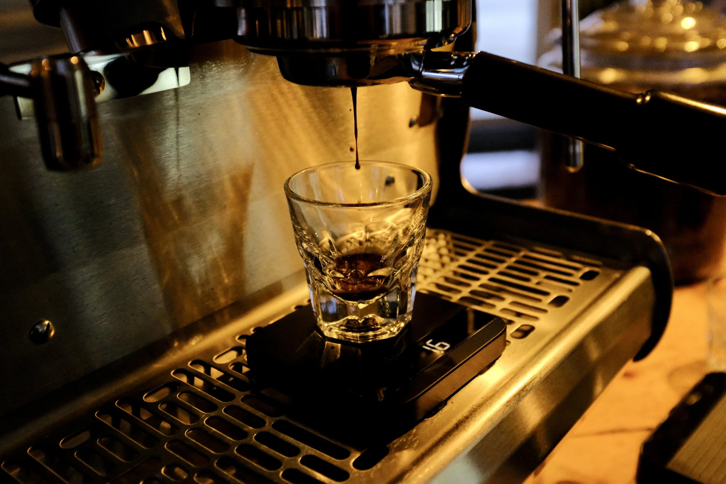 A glass of freshly brewed espresso placed on a coffee machine with a dark coffee drip falling from the machine's spout.