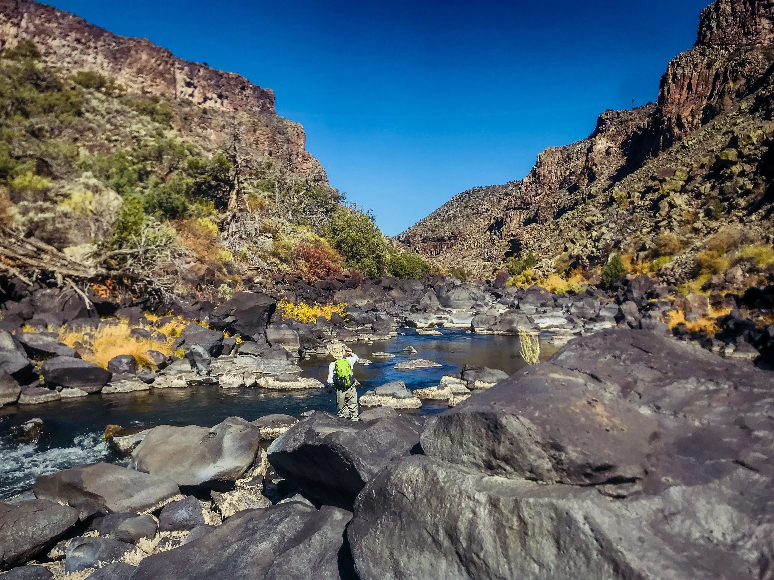Hiker with a bright green backpack stands on rocks by a river in a canyon with colorful autumn foliage, rugged cliffs, and a clear blue sky.