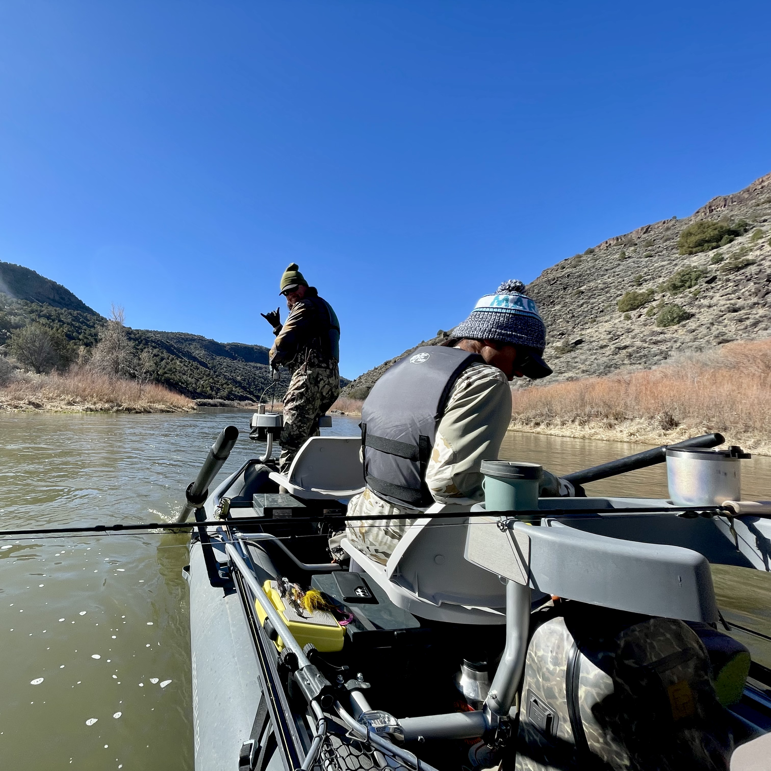 Two people fishing from a small boat on a river with mountainous terrain and clear blue sky in the background.