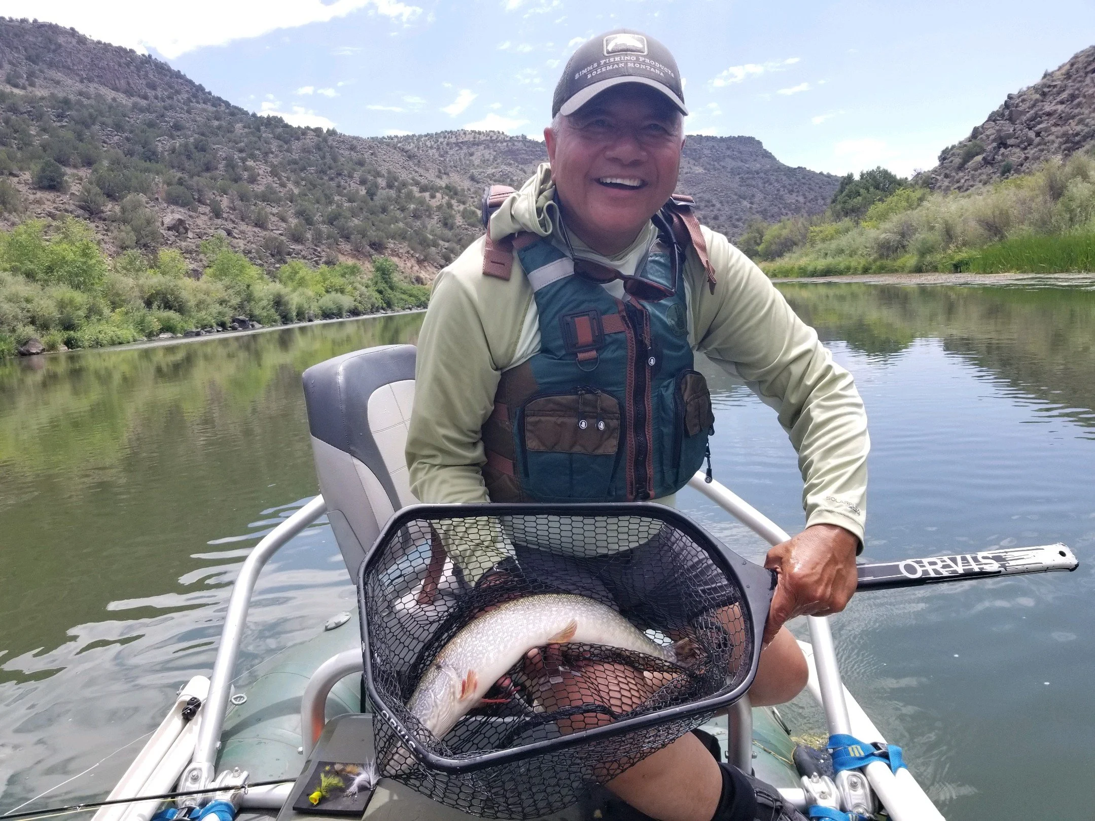 Smiling man in fishing gear in boat on river holding a fish in a net, surrounded by mountains and greenery under a partly cloudy sky.