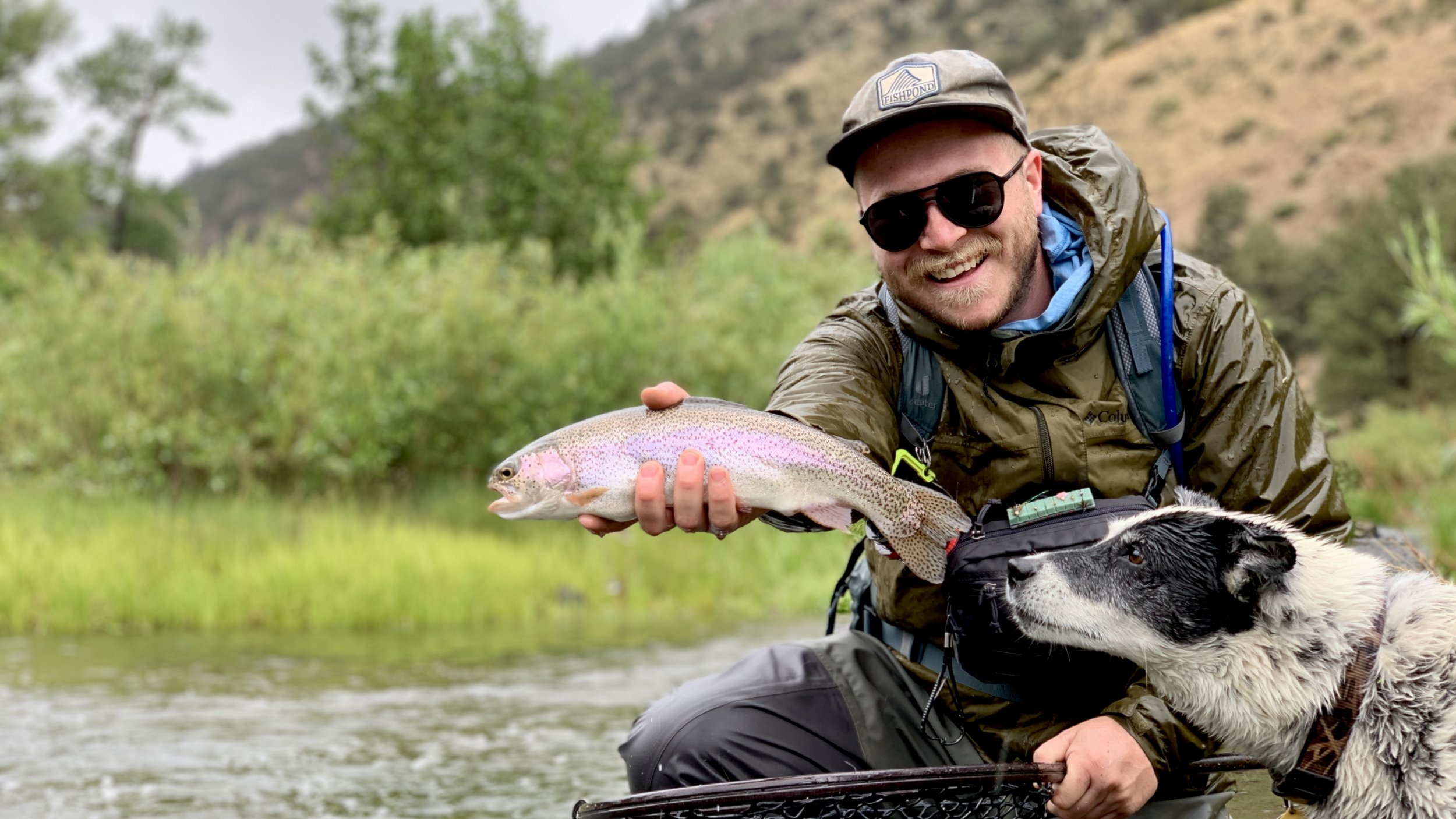 A man with sunglasses, a hat, and a green waterproof jacket holding a rainbow trout, sitting in a river with green trees and hills in the background. A black and white dog is nearby.