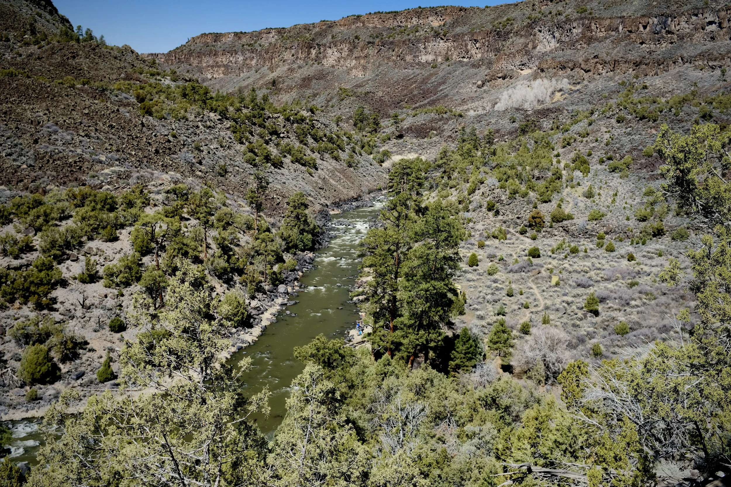 Aerial view of a river flowing through a canyon with rocky, barren hills and green trees.
