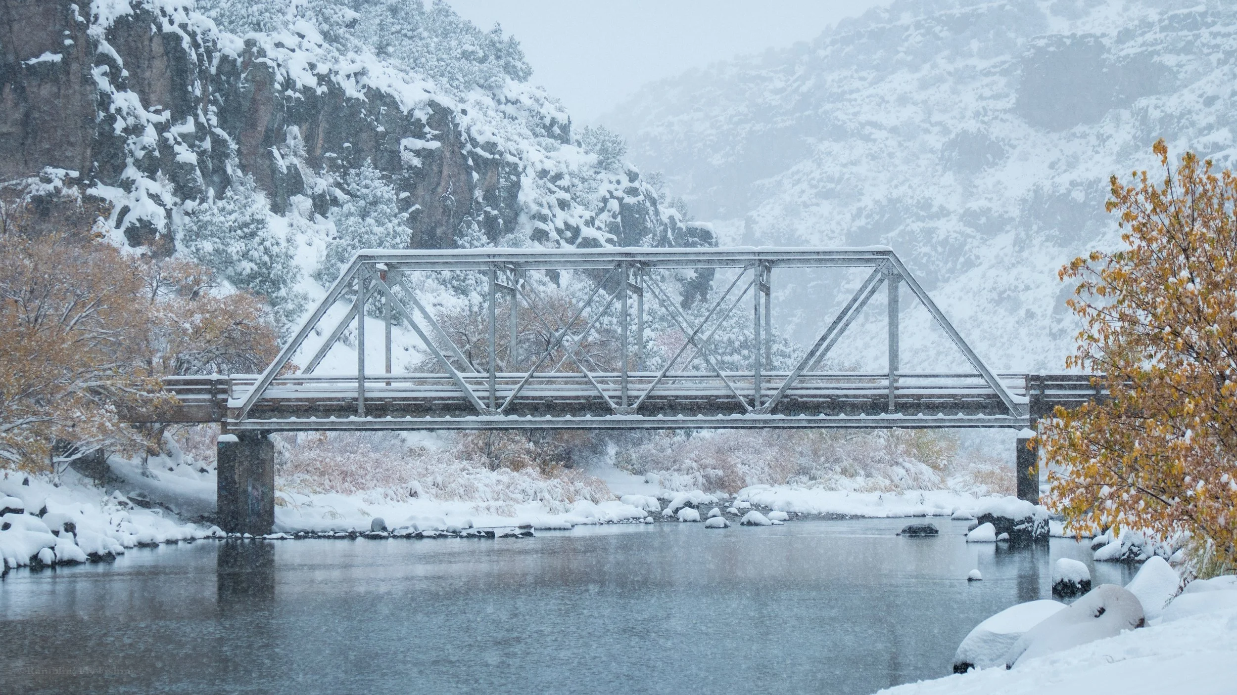 Snow-covered bridge over a river in a mountainous, winter landscape with trees and rocky cliffs.