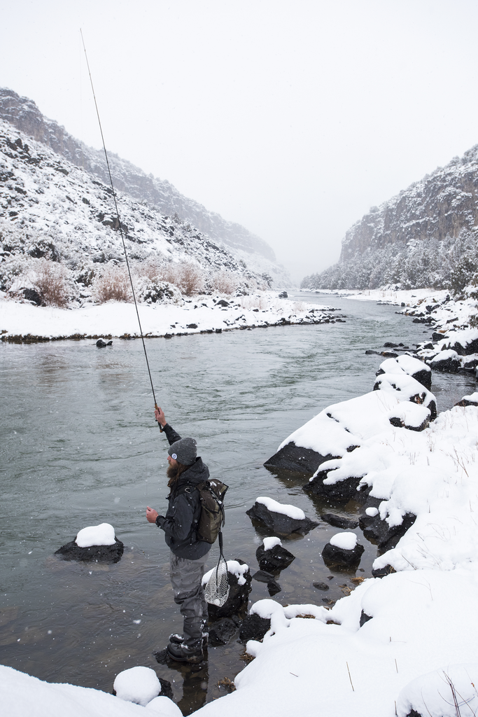 A man fishing in a snow-covered riverbank with mountains in the background.