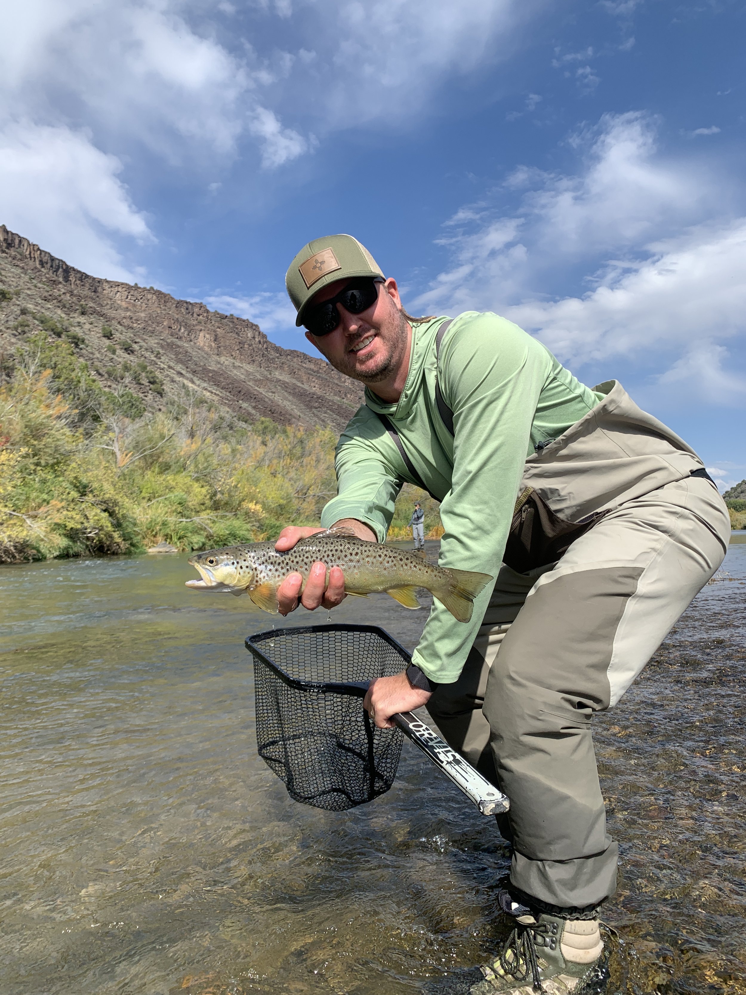 Man holding a freshly caught rainbow trout while standing in a shallow river with a mountain and blue sky in the background.