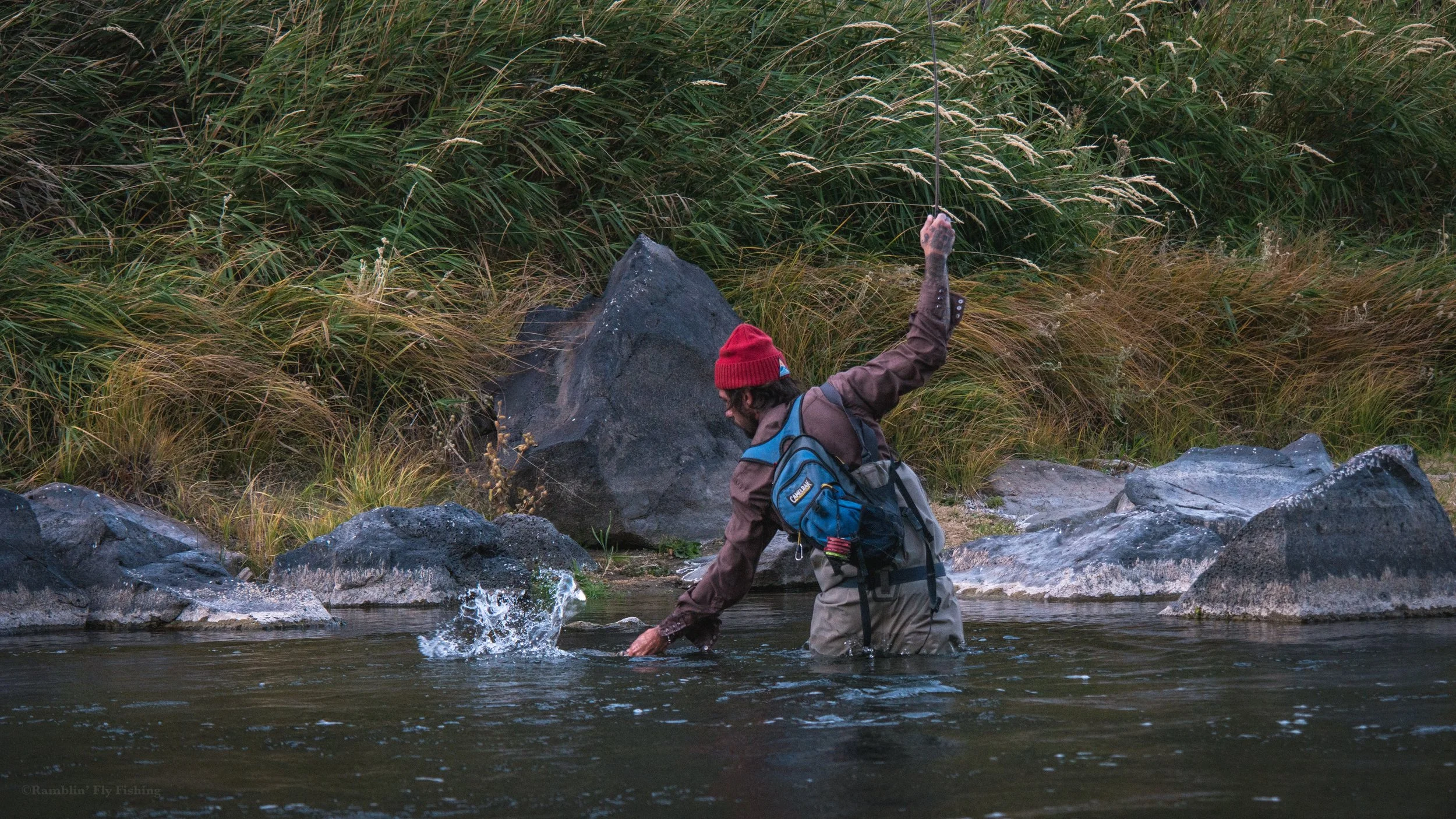A person wearing a red beanie, brown jacket, carrying a blue backpack, wading in a river while fishing with a line and hook, surrounded by rocks and tall grass.