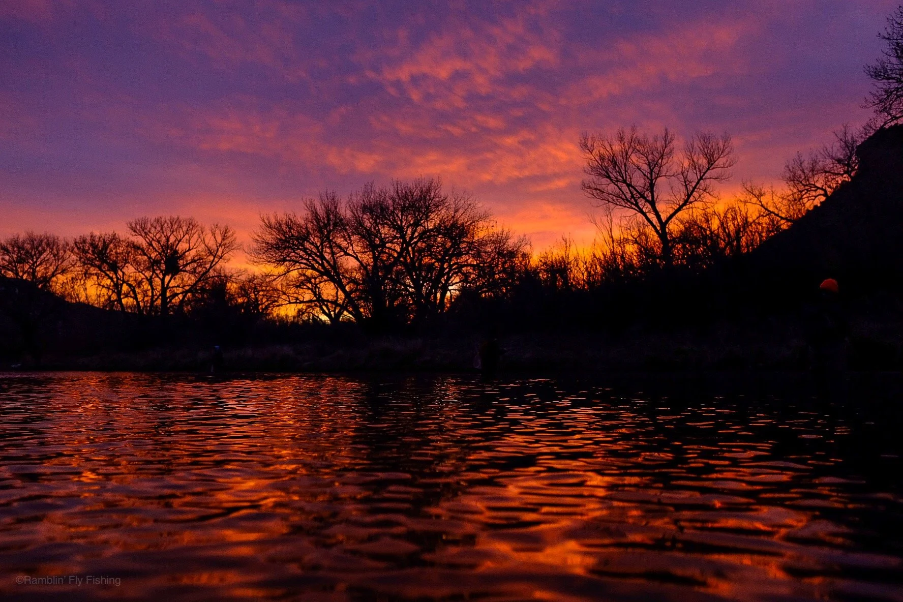Sunset over a river with silhouetted trees and hills, vibrant orange, purple, and pink sky reflections on the water.