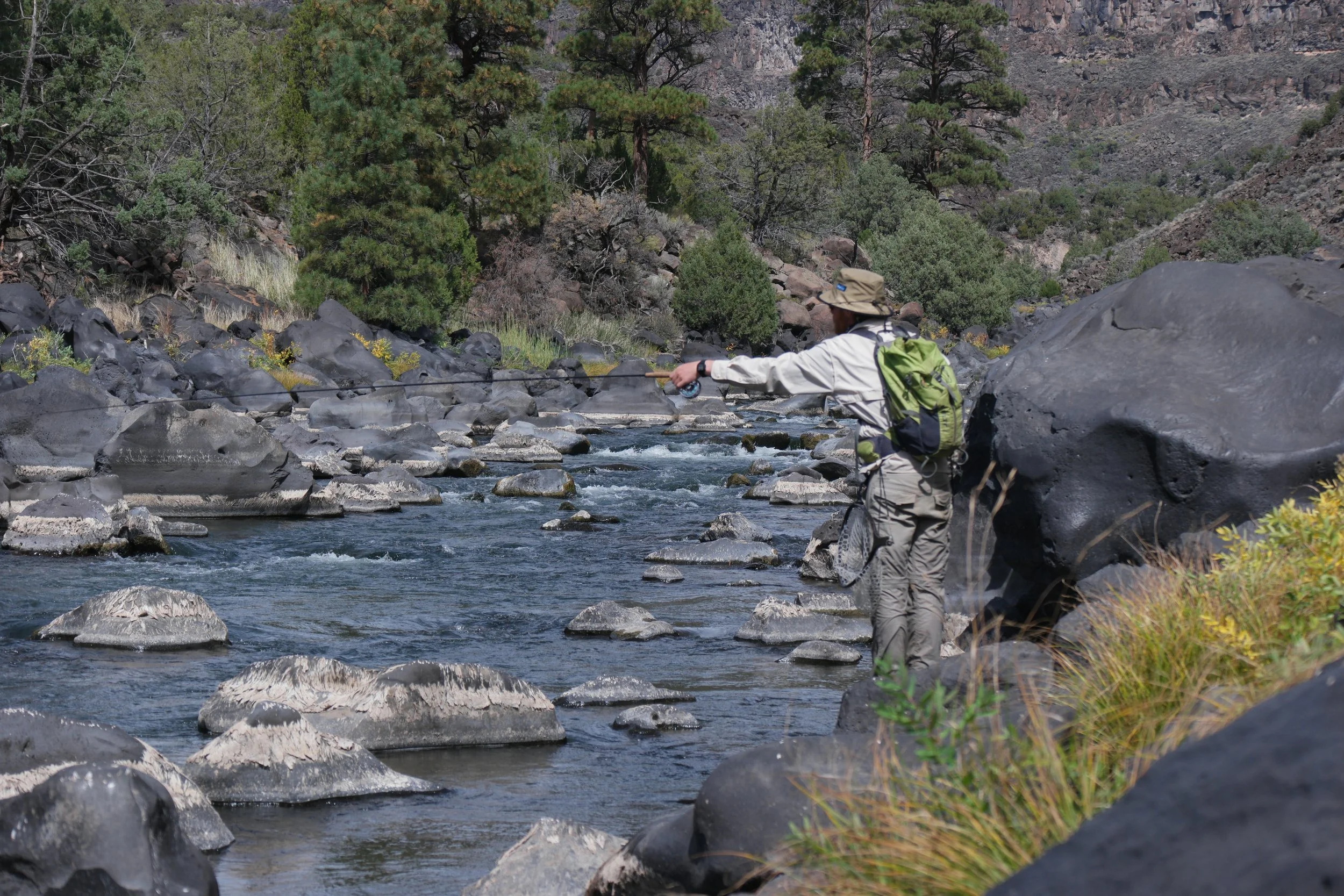 A person fishing in a river surrounded by rocks and trees in a mountainous landscape.