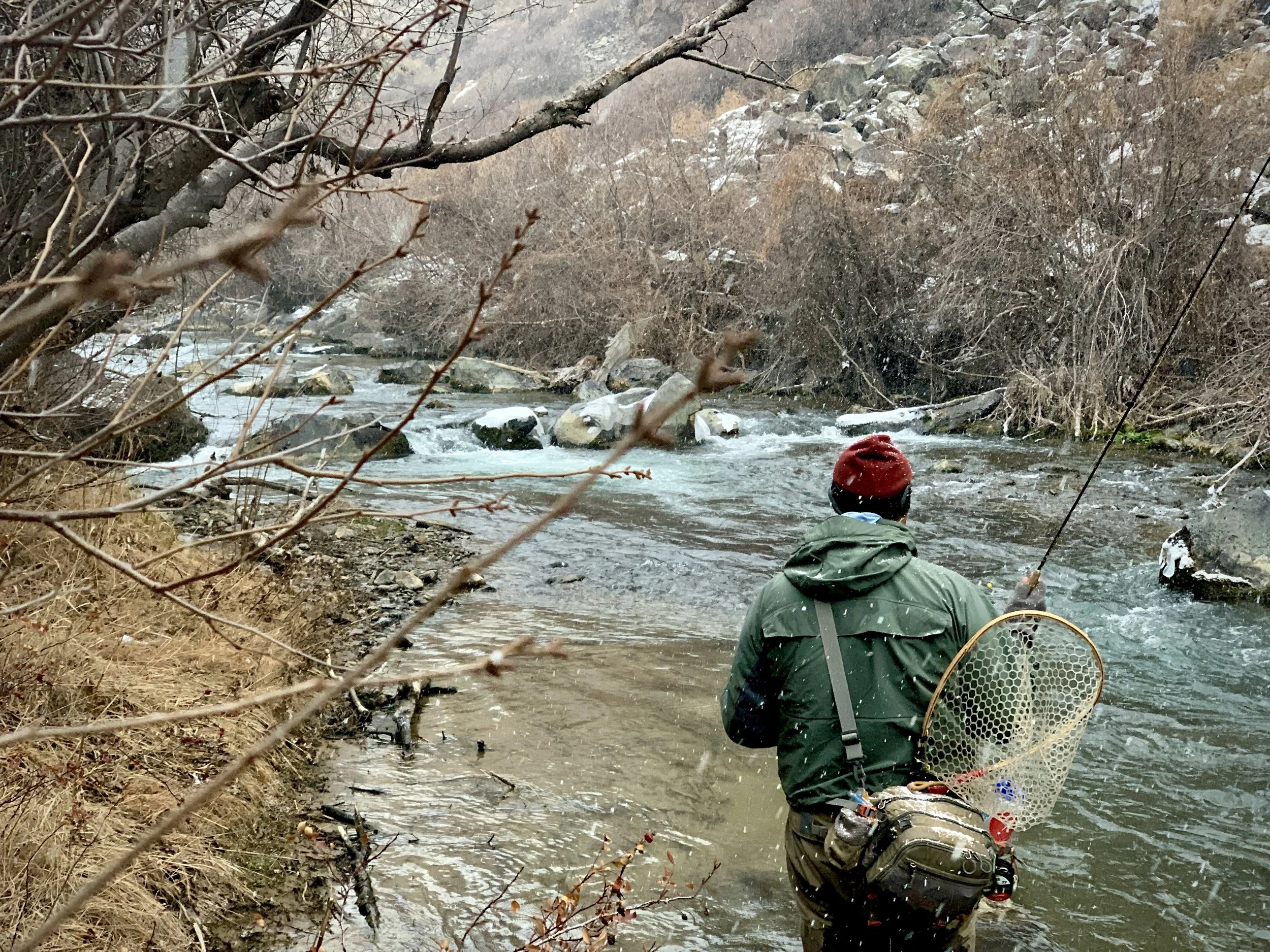 A person standing in a river with a fishing rod, wearing a red beanie and a green jacket, surrounded by leafless bushes and rocks in a cold, mountainous environment.