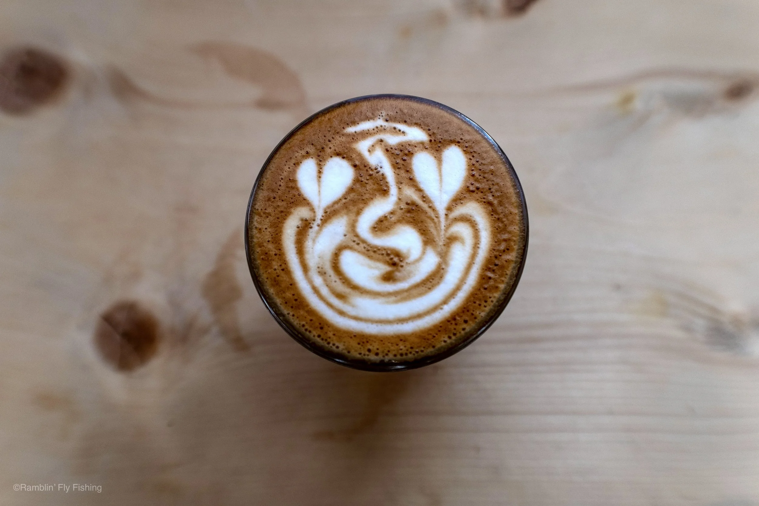 Top-down view of a latte in a glass with foam art resembling a floral pattern on top, on a light wooden surface.
