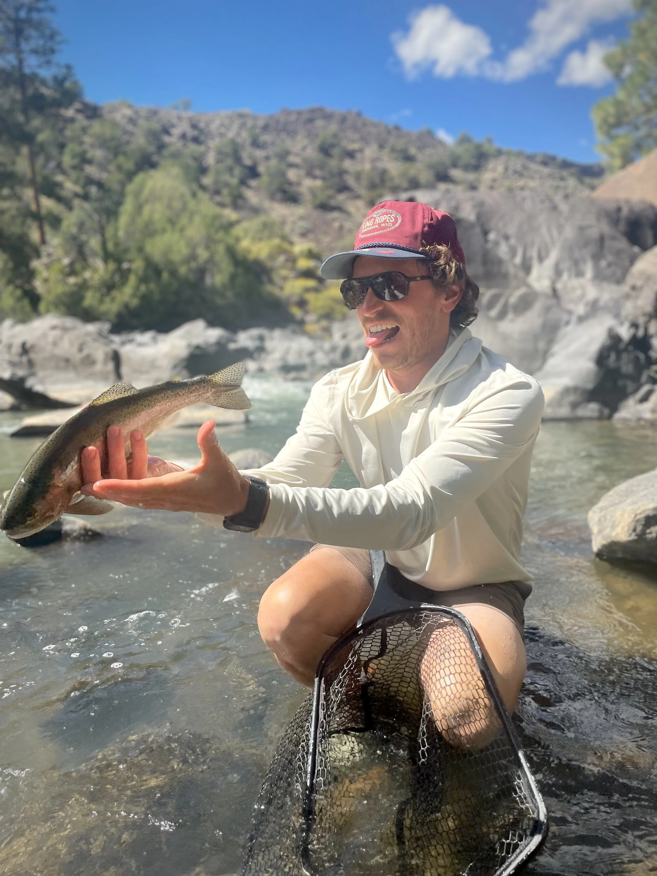 A man kneeling in a river, holding a fish and displaying it. He is wearing sunglasses, a hat, a long-sleeve shirt, and shorts, with a fishing net on his lap. The background features rocky terrain and a clear blue sky with some clouds.
