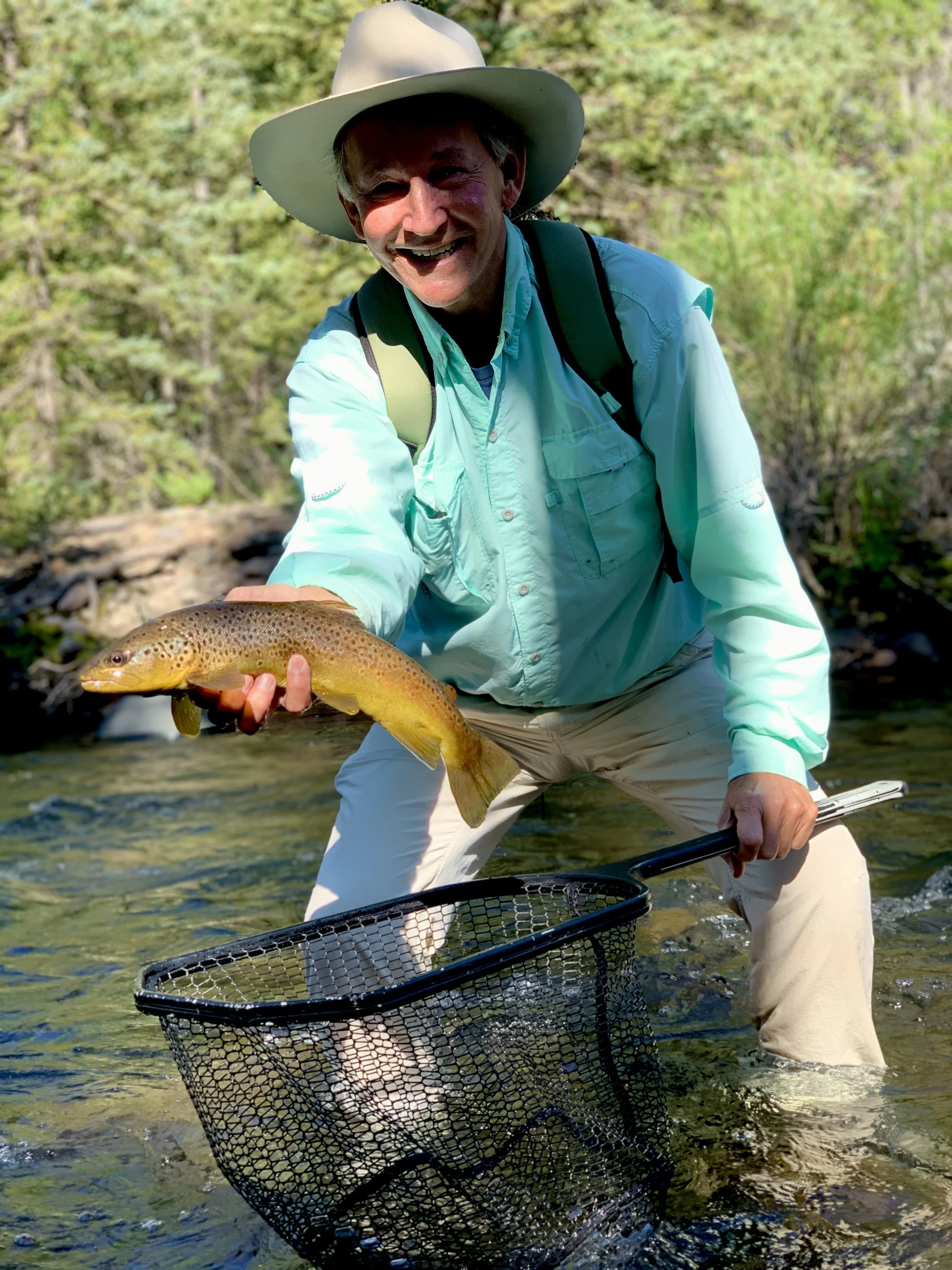 A person in outdoor clothing and a wide-brimmed hat holding a fish over a stream, with a fishing net in one hand and smiling at the camera.