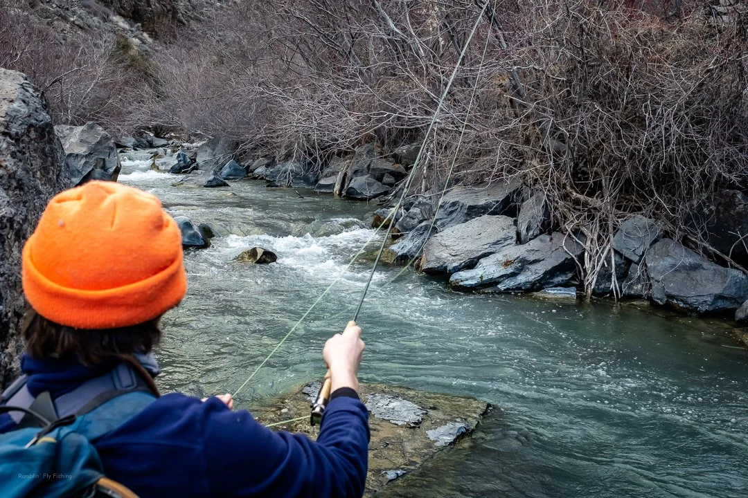 A person wearing an orange beanie and a blue jacket fishing in a river with rocks and bare trees on the riverbank.