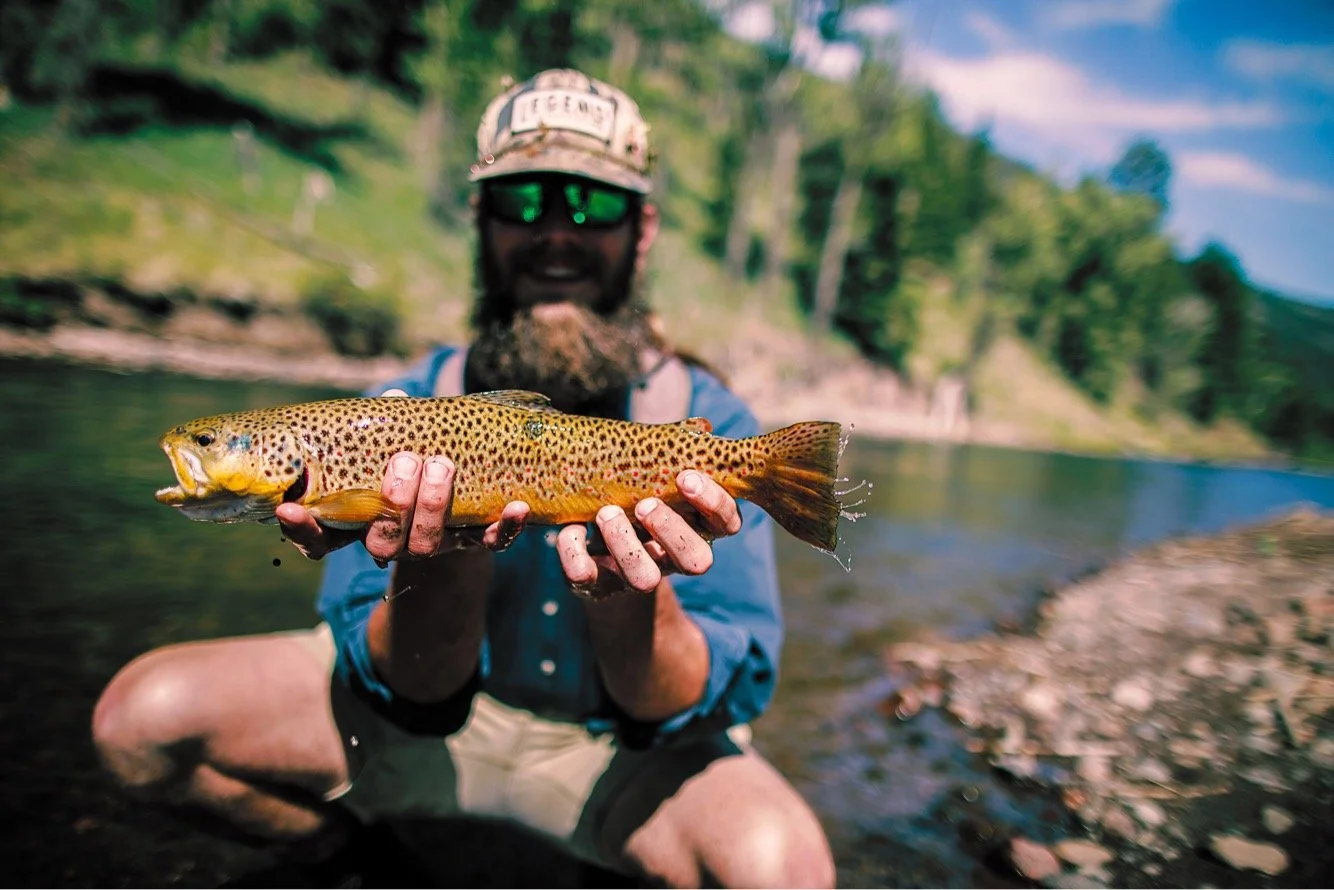 Man with a beard, wearing sunglasses, a cap, and outdoor clothing, holding a freshly caught colorful fish near a river with trees and mountains in the background.