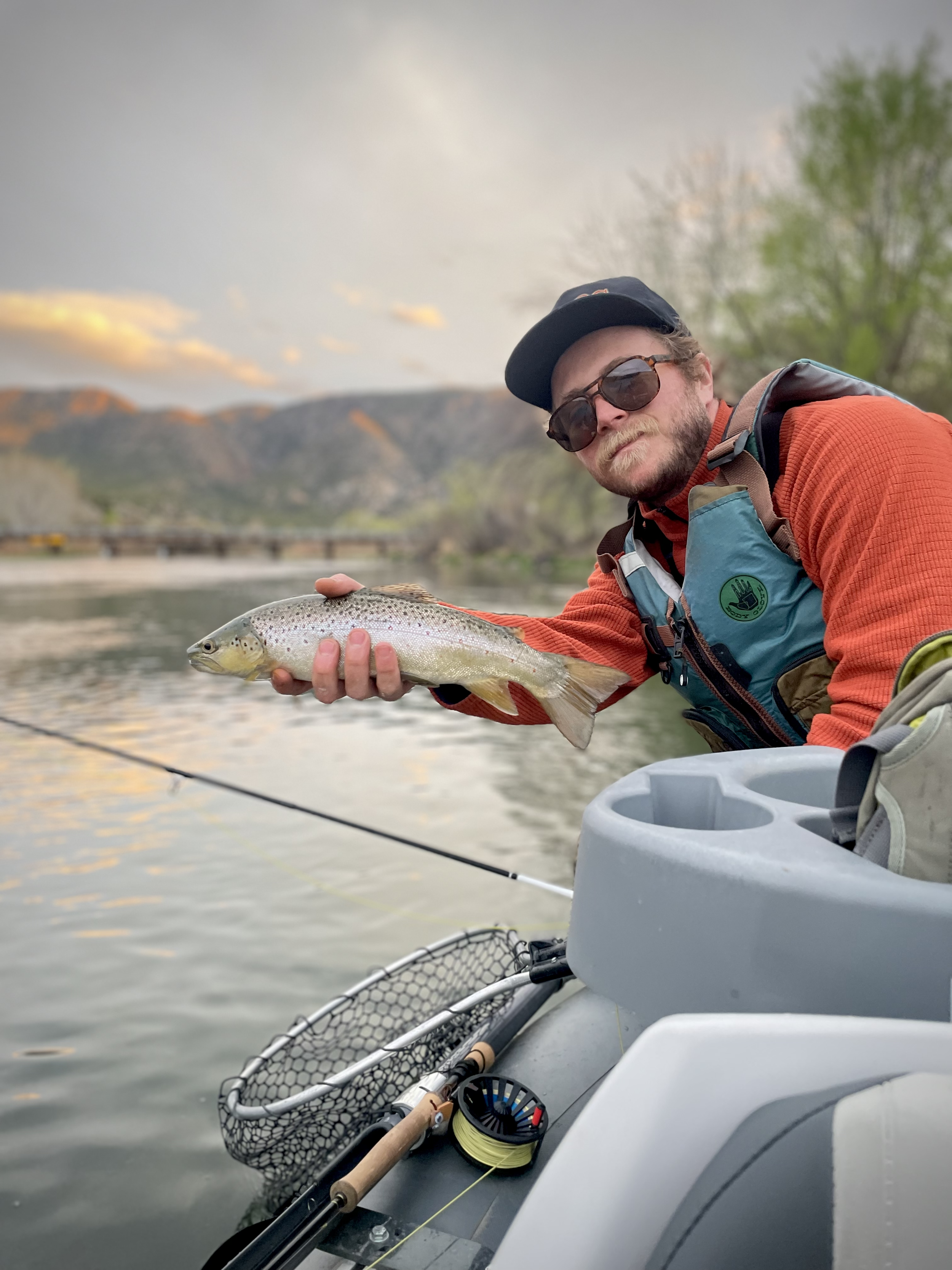 Man in sunglasses holding a fish on a boat with fishing gear and a scenic mountain and lake background during sunset.