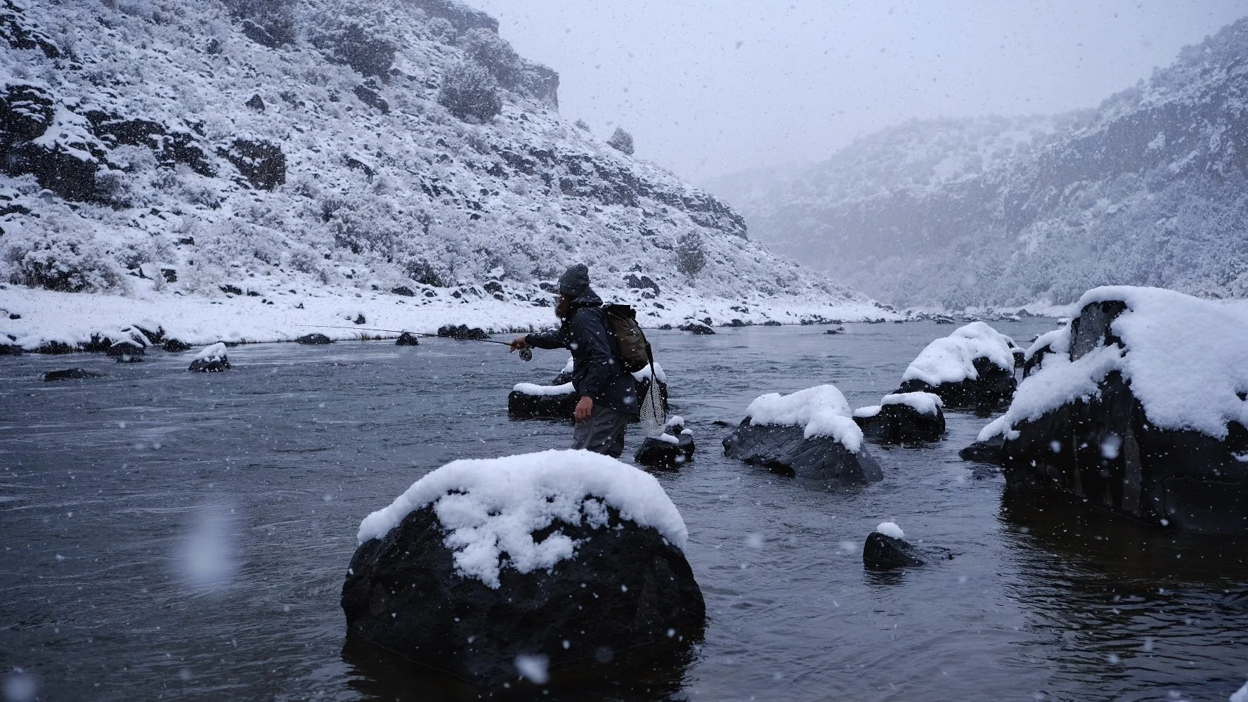 A person fly fishing in a snowy river surrounded by snow-covered rocks and mountains, with snow falling.