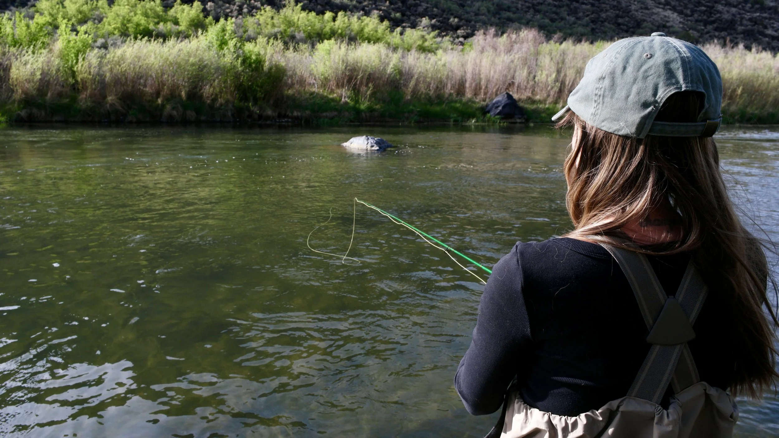 A woman with long hair wearing a gray cap and black jacket fishing in a river surrounded by green trees and shrubs.