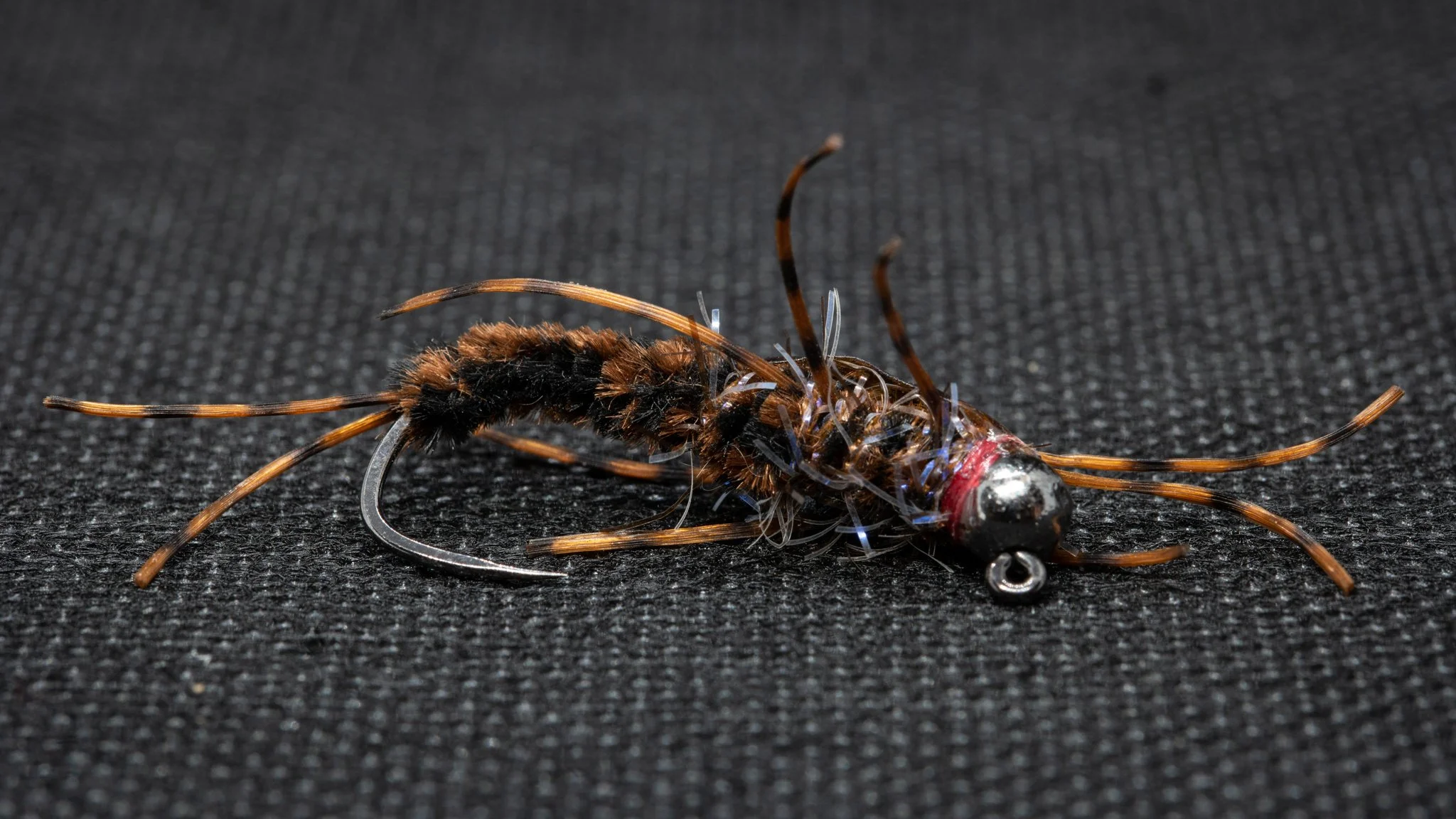Close-up of a fishing lure resembling a small insect, with a hook, black and brown fuzzy body, and thin antennae on a textured black surface.